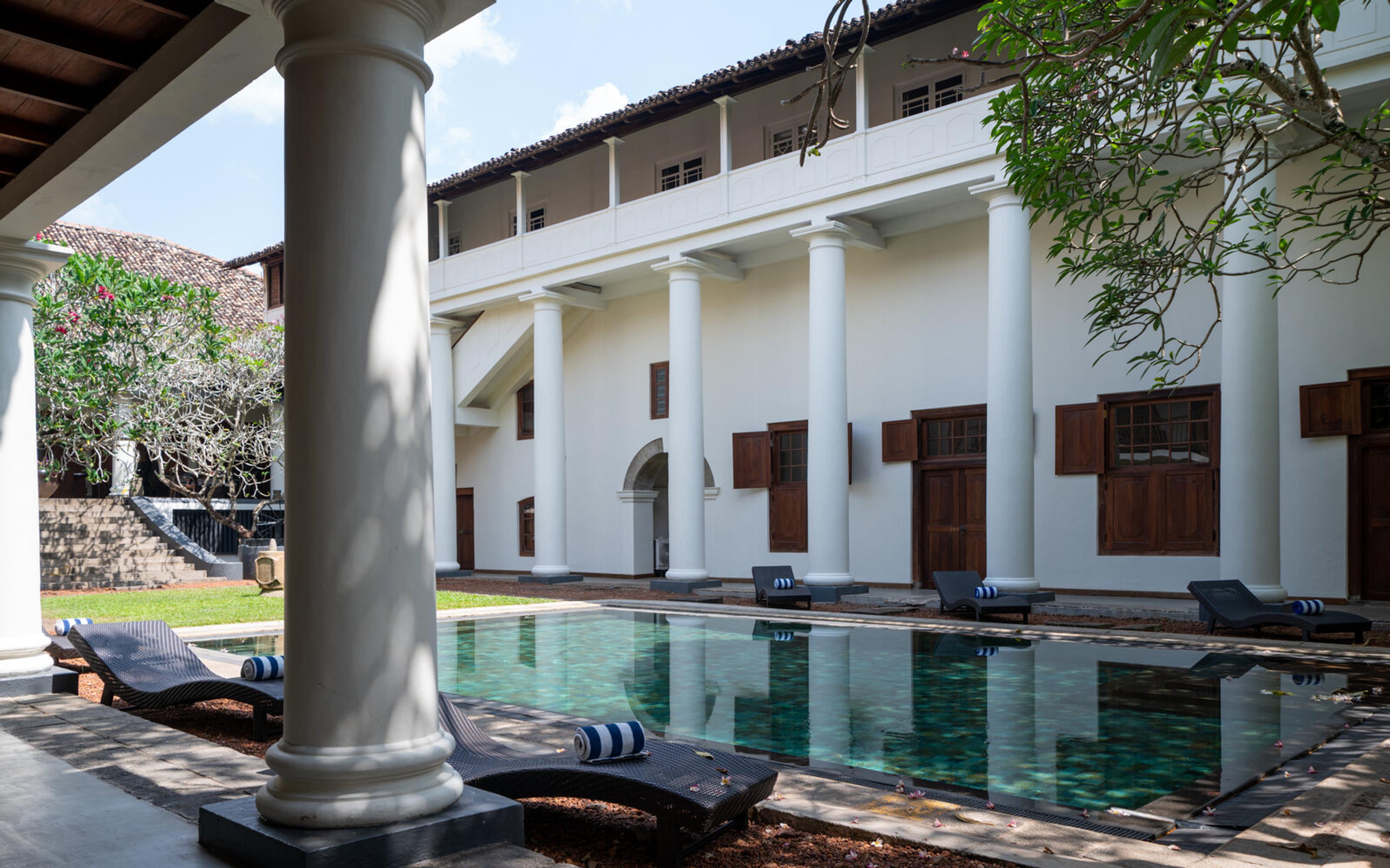 A pool along the side of a white colonnaded building with shuttered doors, seen from under a veranda with loungers set along the water's edge.
