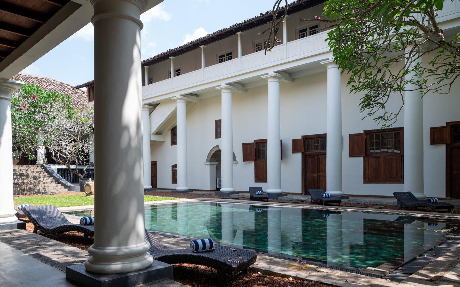 A pool along the side of a white colonnaded building with shuttered doors, seen from under a veranda with loungers set along the water's edge.