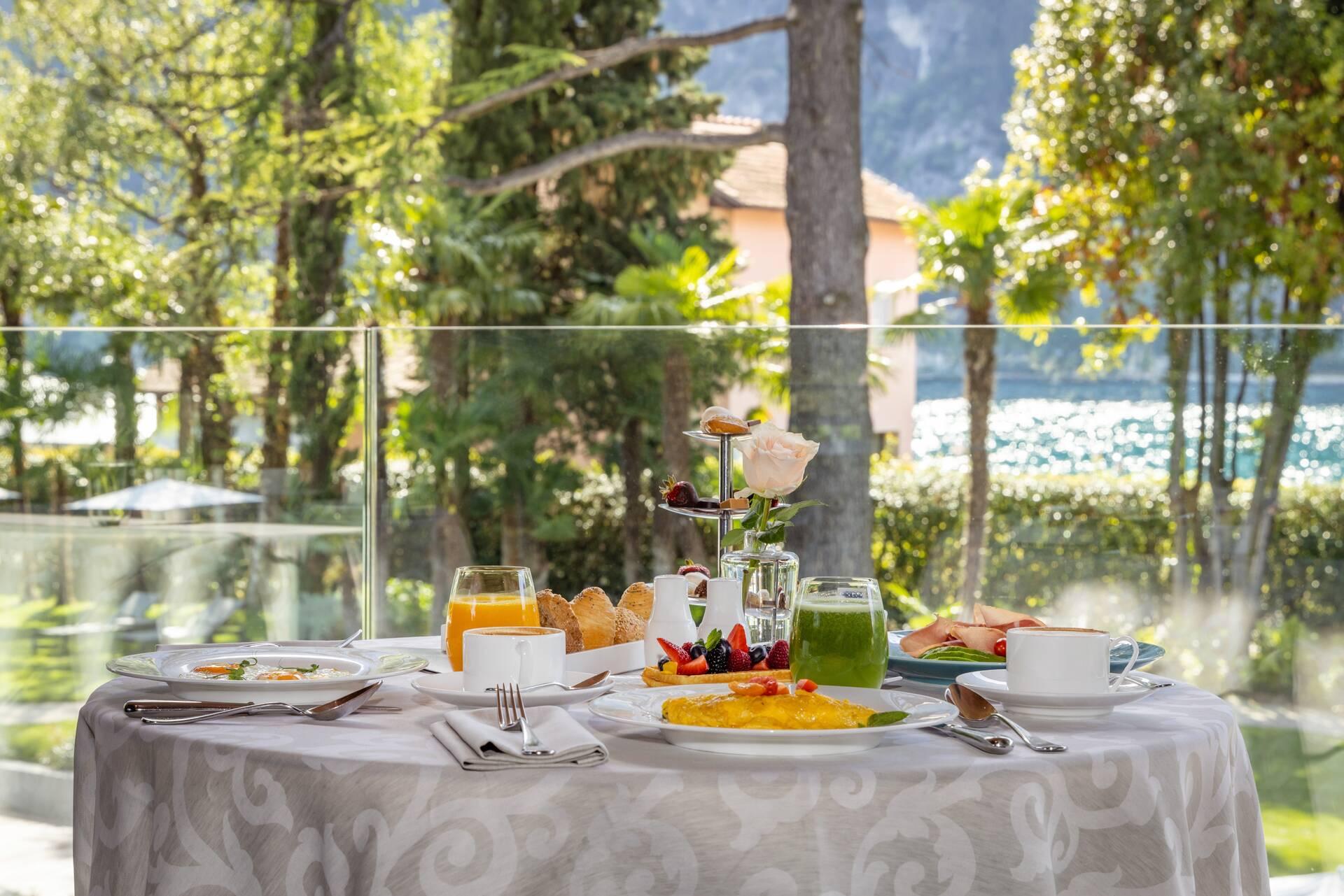 A table set for breakfast on a terrace, with a plate of eggs, juice, pastries and fruit, and a lake visible through the trees beyond.