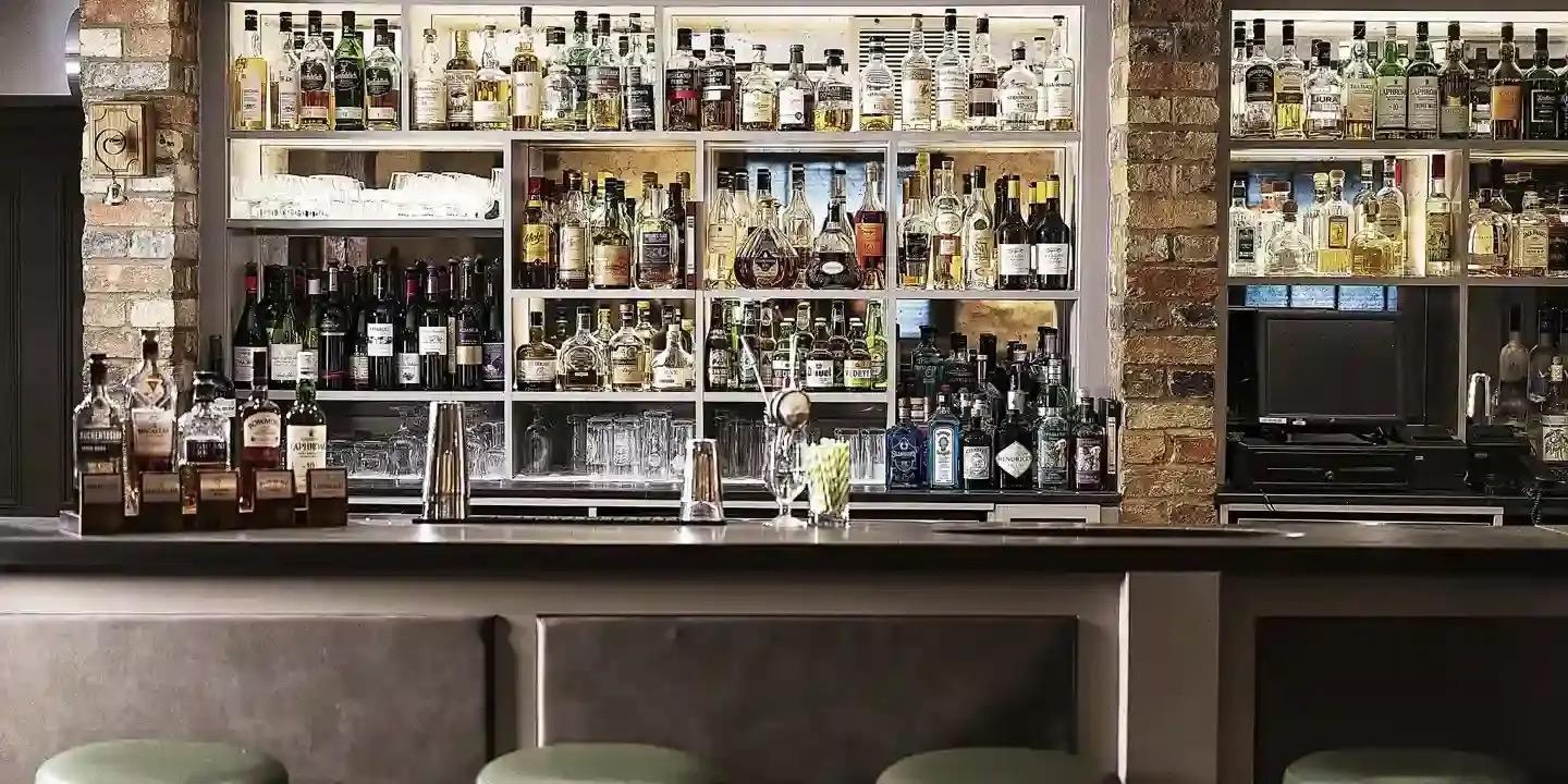 A bar with glass-fronted shelves of spirits and wine bottles on a back wall with brick pillars at each end, a dark counter and green-topped stools in front.