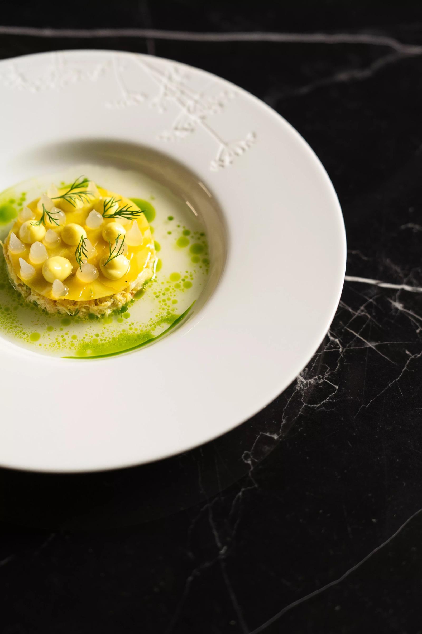 A pale plated dish topped with small yellow spheres and herb oil sits on a white plate on a dark marble table.