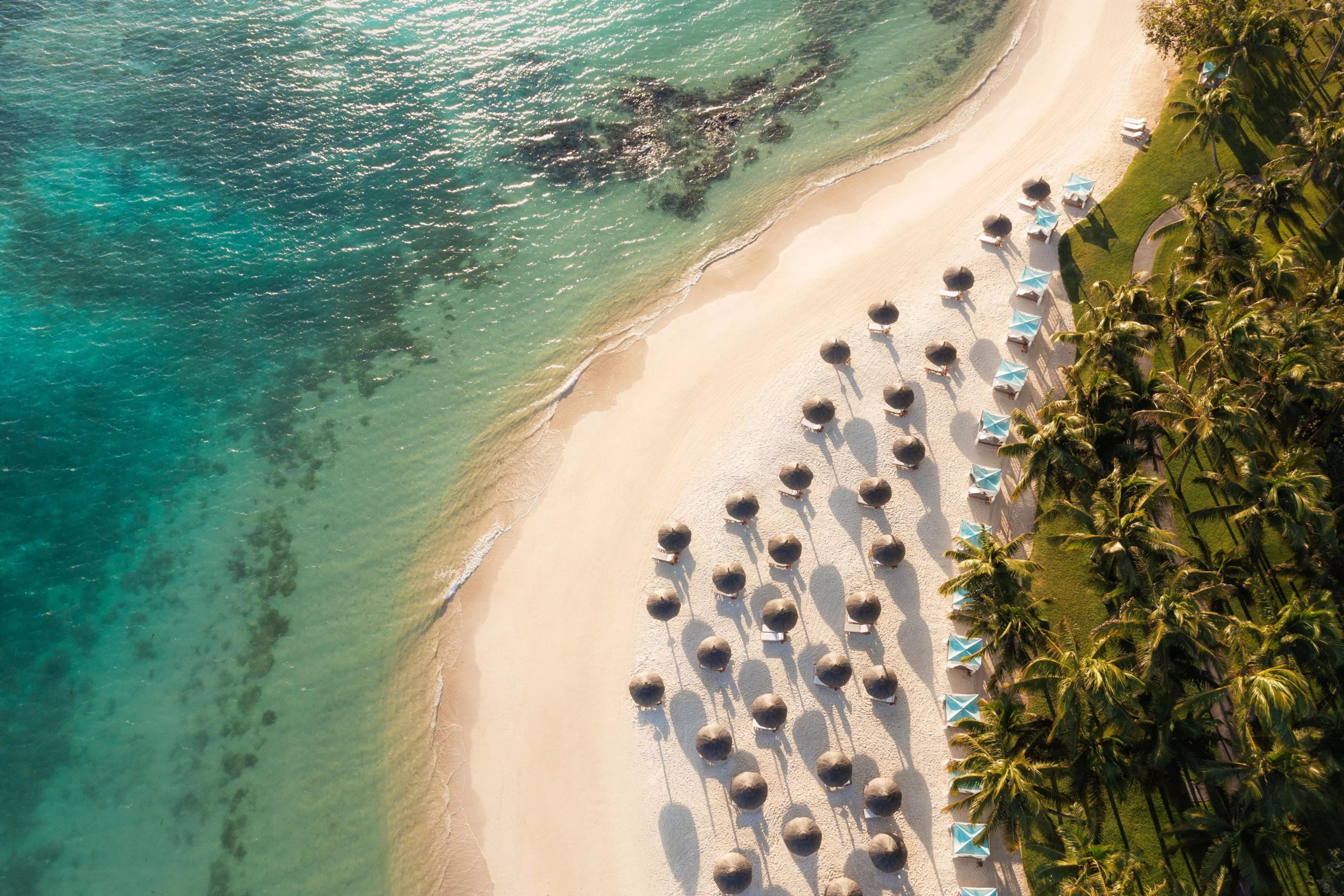 An aerial view of a curving white sand beach lined with rows of thatched umbrellas and sun loungers beside turquoise water.