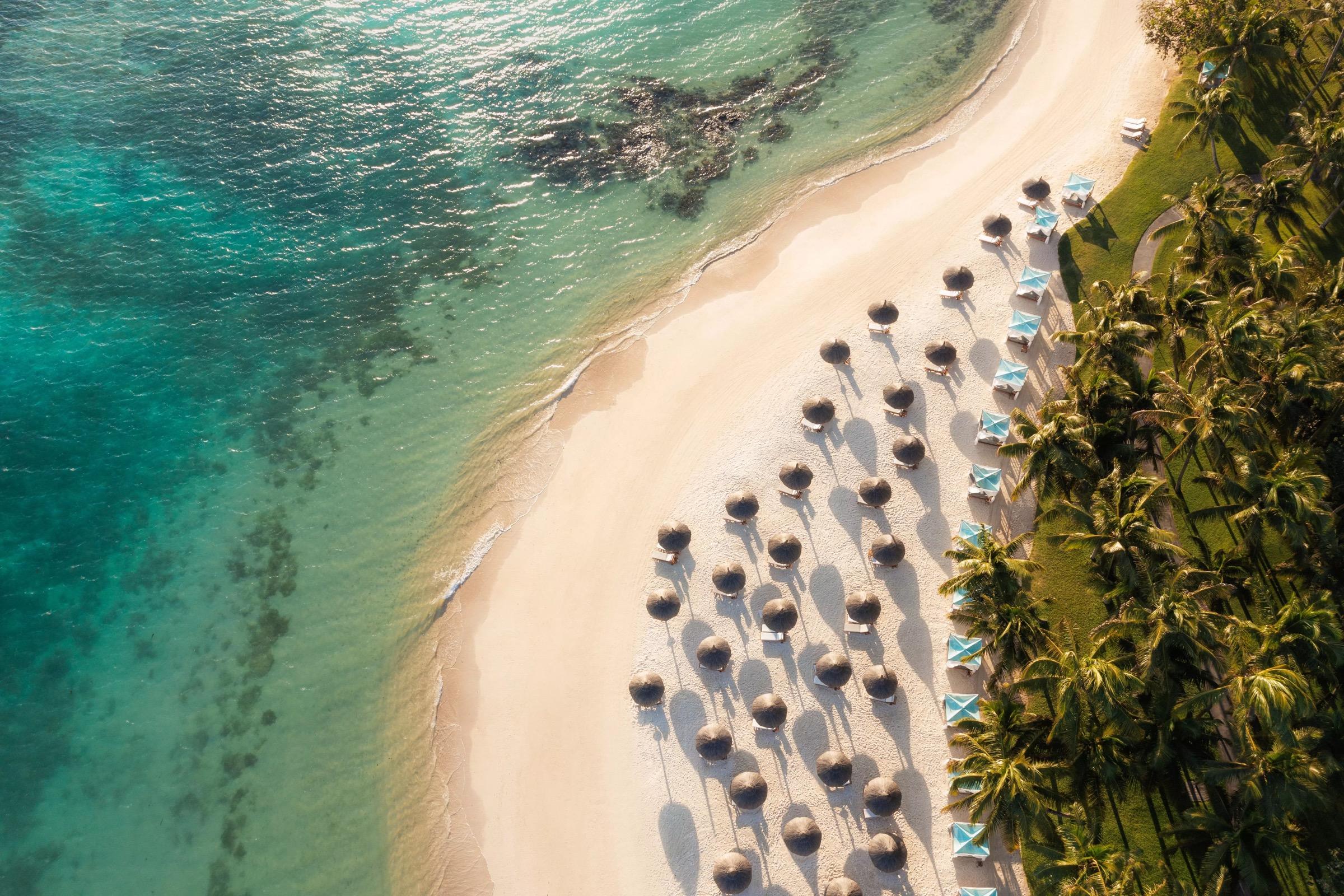 An aerial view of a curving white sand beach lined with rows of thatched umbrellas and sun loungers beside turquoise water.