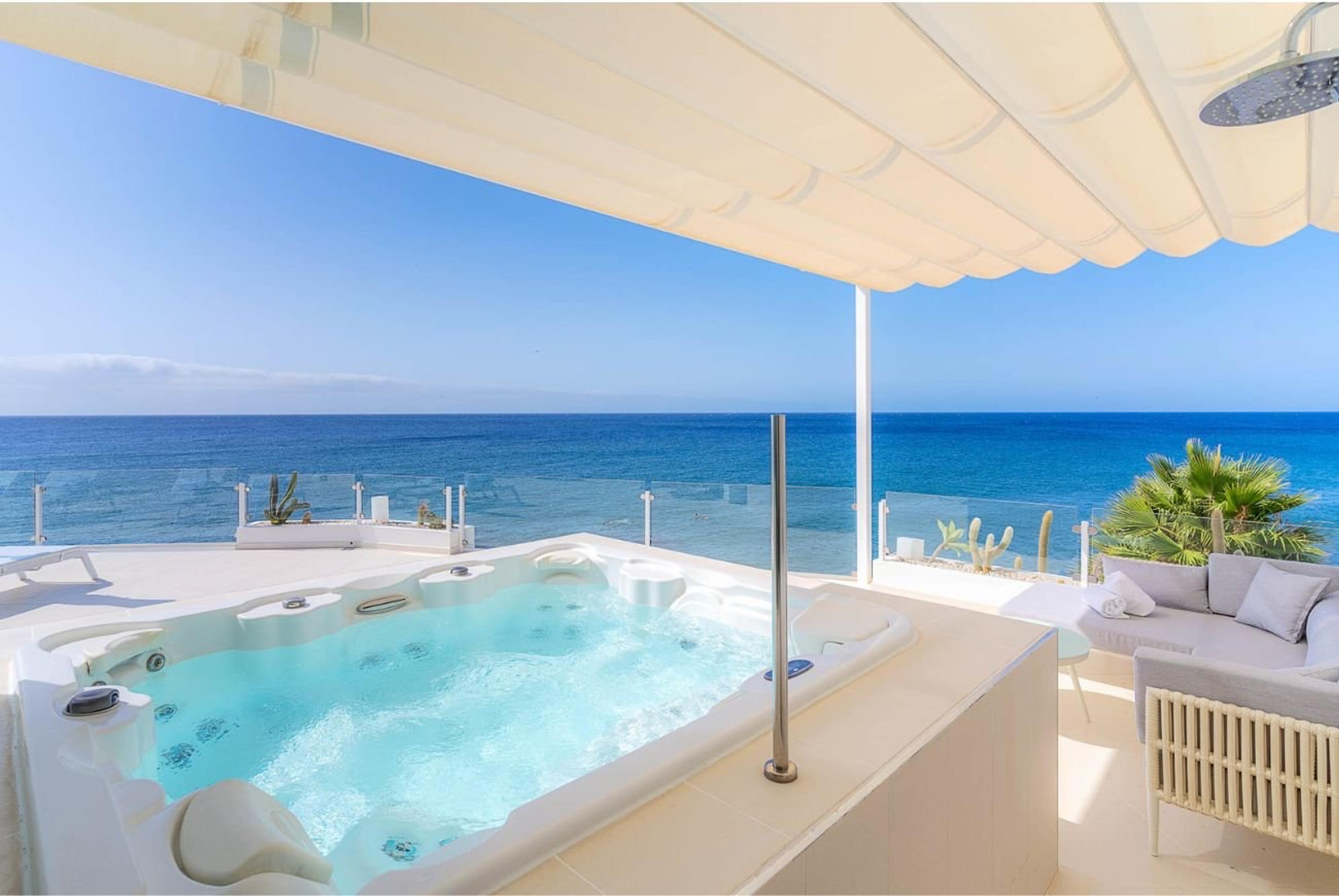 A private outdoor jacuzzi on a terrace under a canopy, with the sea filling the view and a lounge seating area to the side.