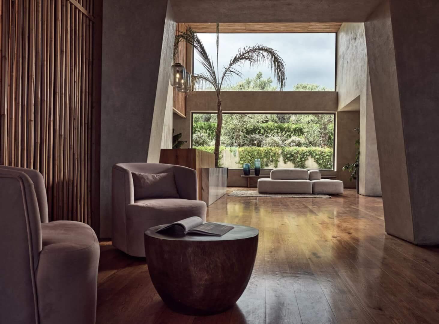 A sitting area with low armchairs and a round table, framed by a bamboo screen and a tall window onto a garden.