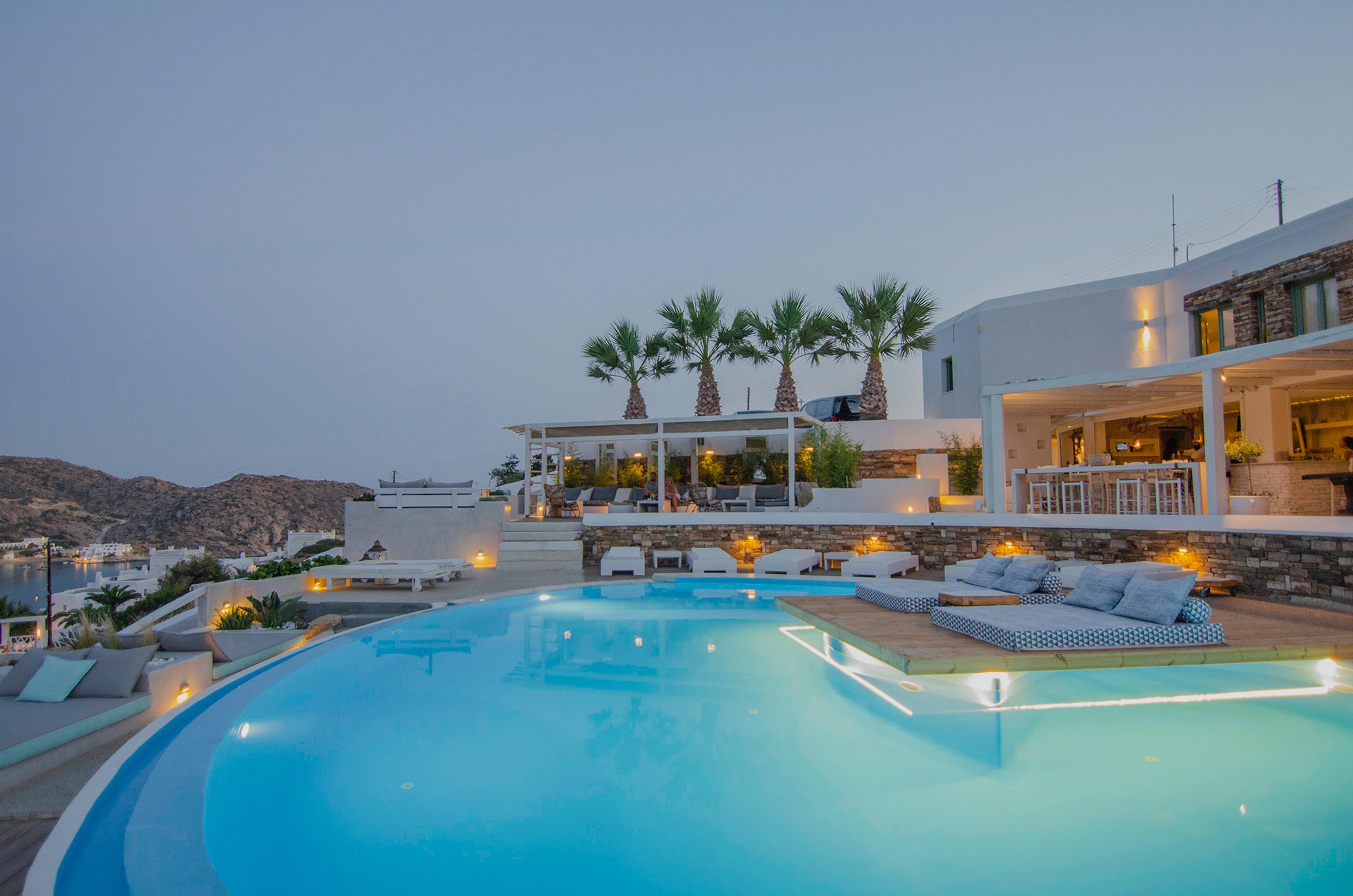 A pool at dusk with its lights on, white loungers and palm trees on the terrace, and the hotel's bar and building lit on the right.