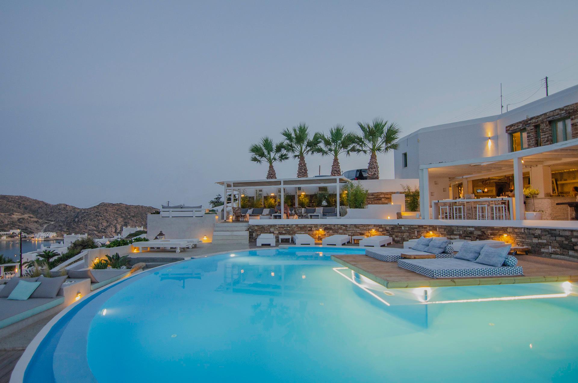 A pool at dusk with its lights on, white loungers and palm trees on the terrace, and the hotel's bar and building lit on the right.