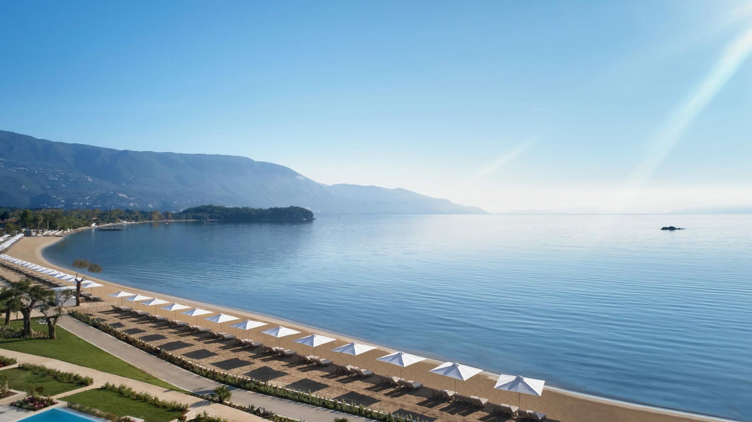 An aerial view of a sandy beach lined with white umbrellas and sunbeds beside calm sea, with mountains in the distance.