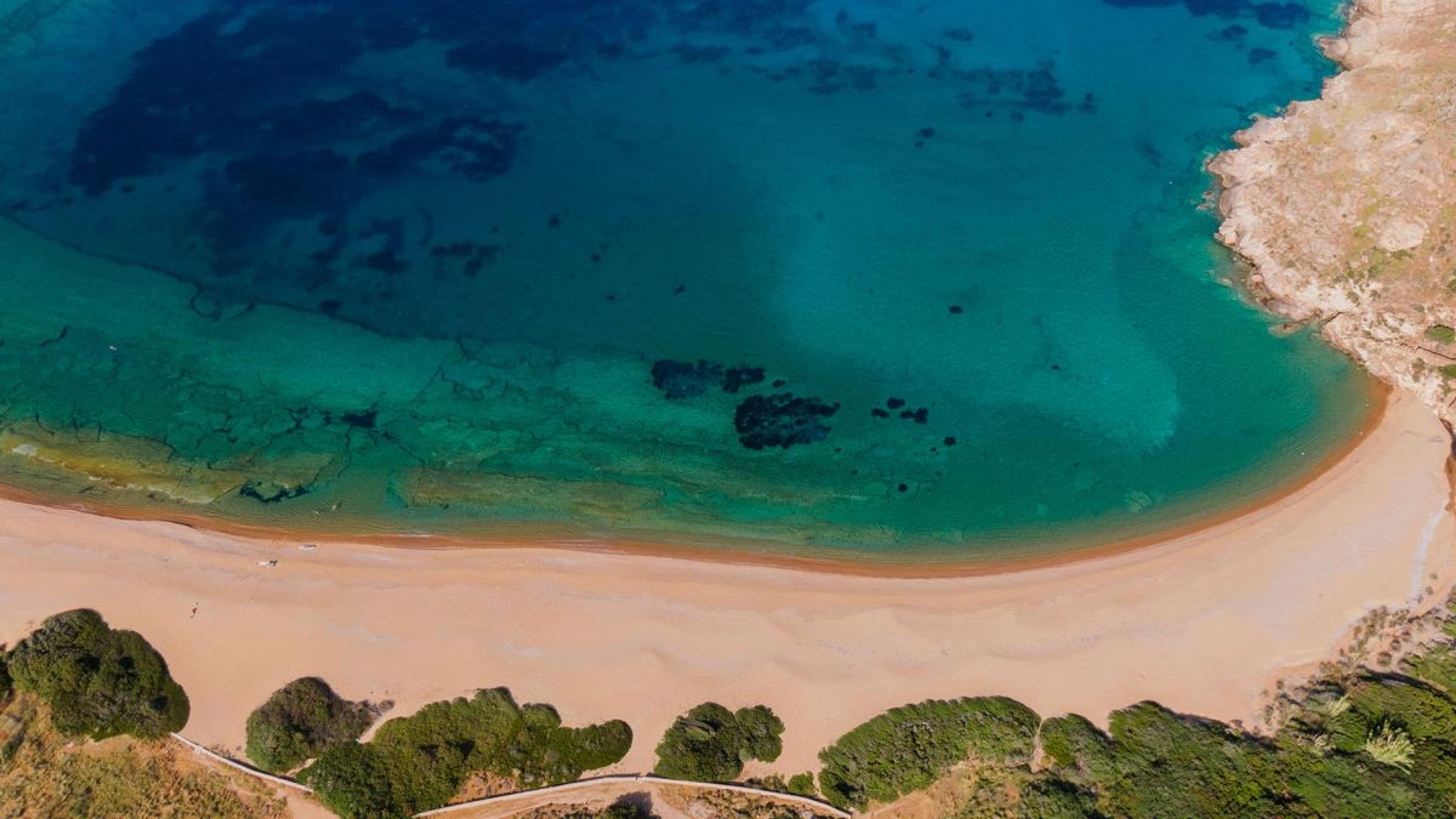 Aerial view of a sandy cove with clear green water, dark rock patches on the seabed, and scrub on the headland at the bottom edge.
