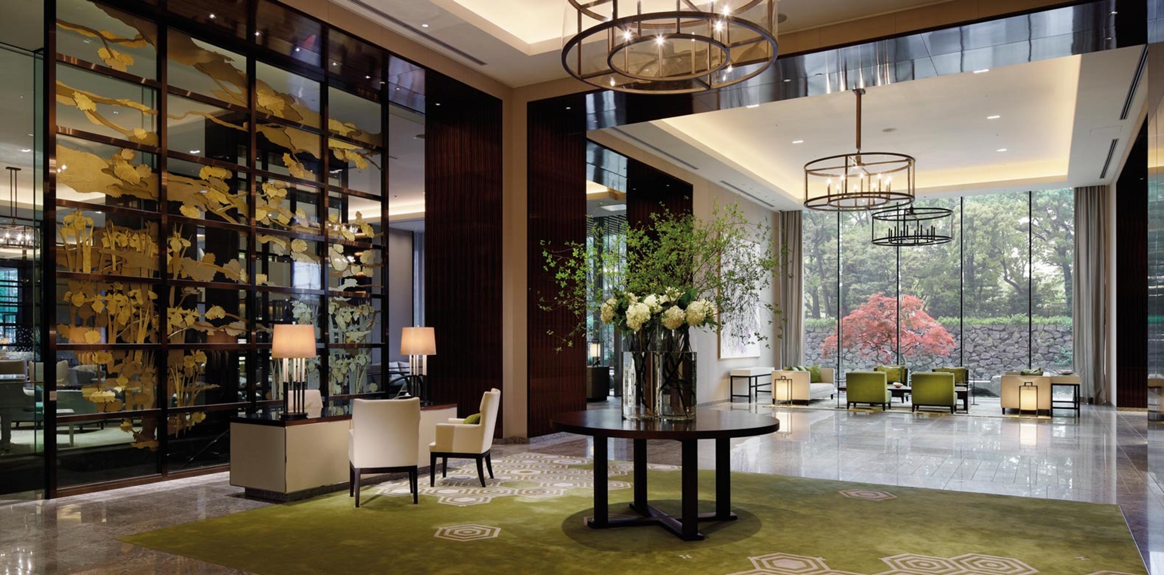 A hotel lobby with a gold leaf screen, a round table of white flowers, drum chandeliers and a lounge beyond glass walls with a red maple tree outside.