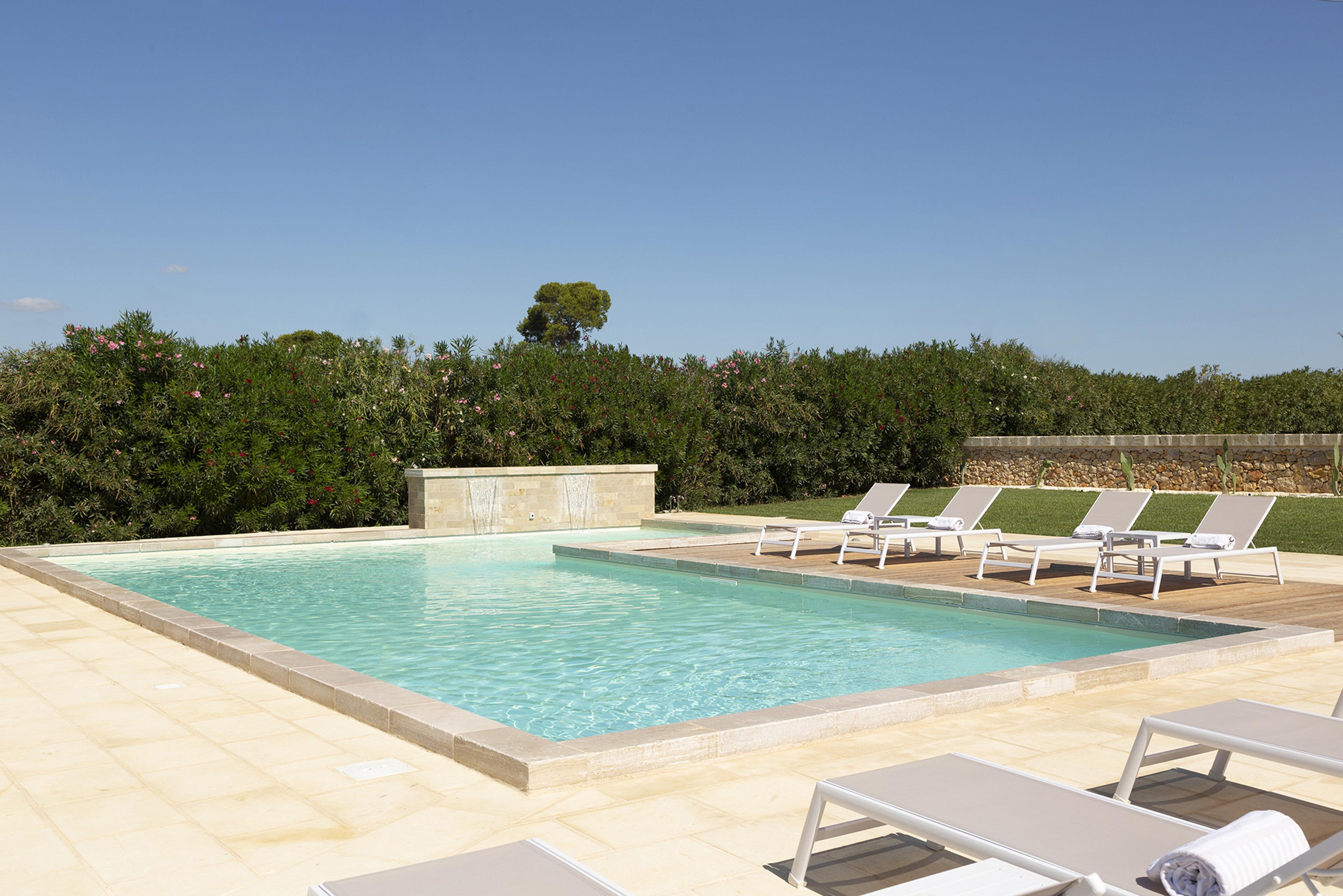 An empty rectangular pool bordered by lawn and shrubs, with sunbeds and a water feature at one end.