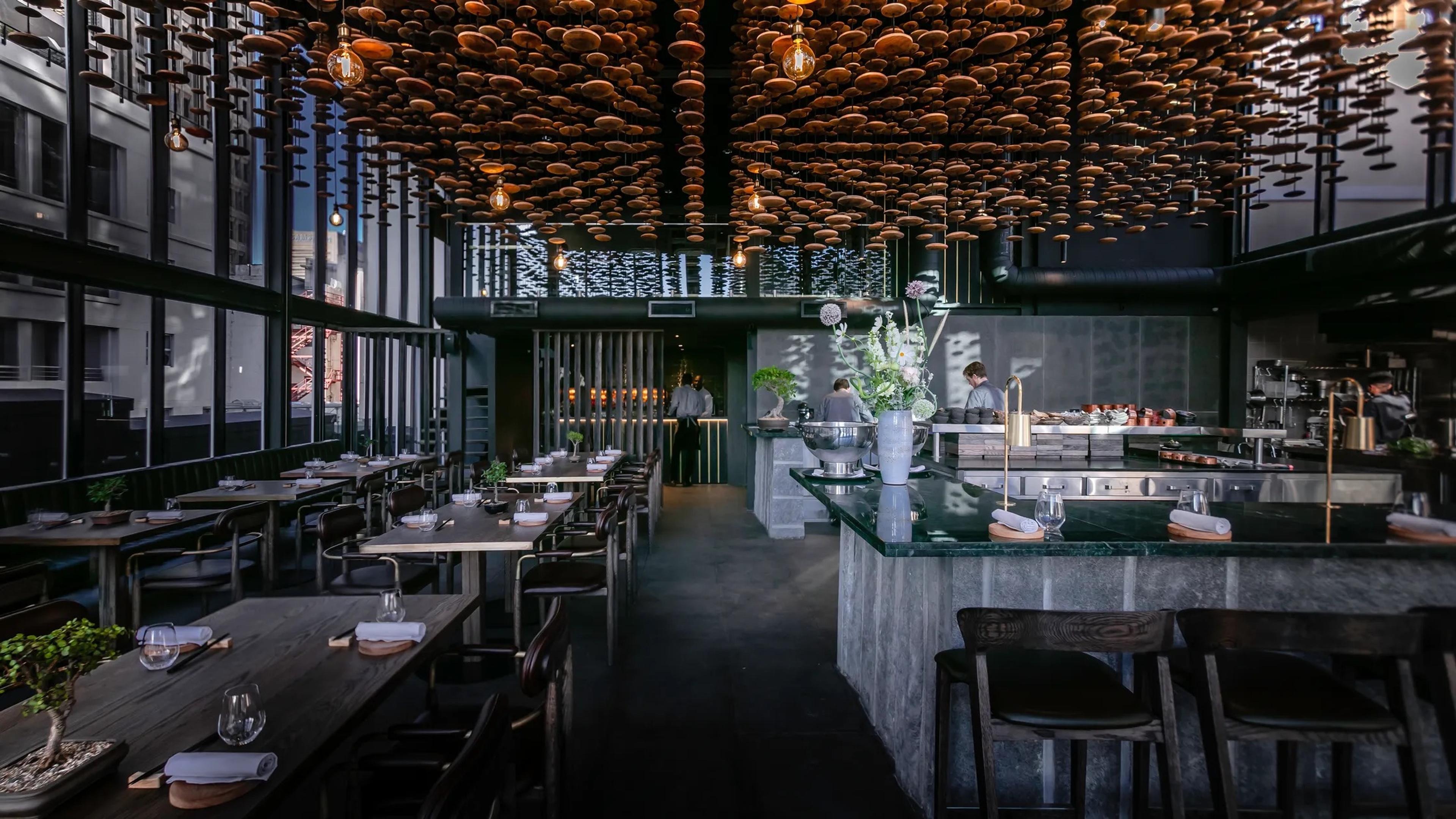 A dark dining room of wooden tables under a ceiling of folded paper forms, with an open kitchen and bar at the far end