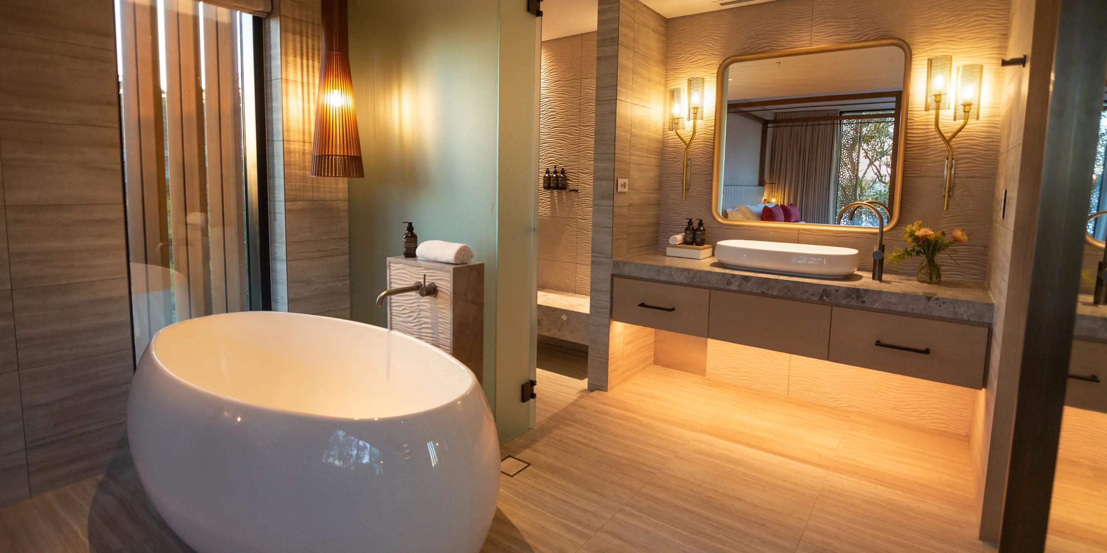 A freestanding oval bathtub in front of a double vanity, with a walk-in shower and a bedroom reflected in the mirror behind it.