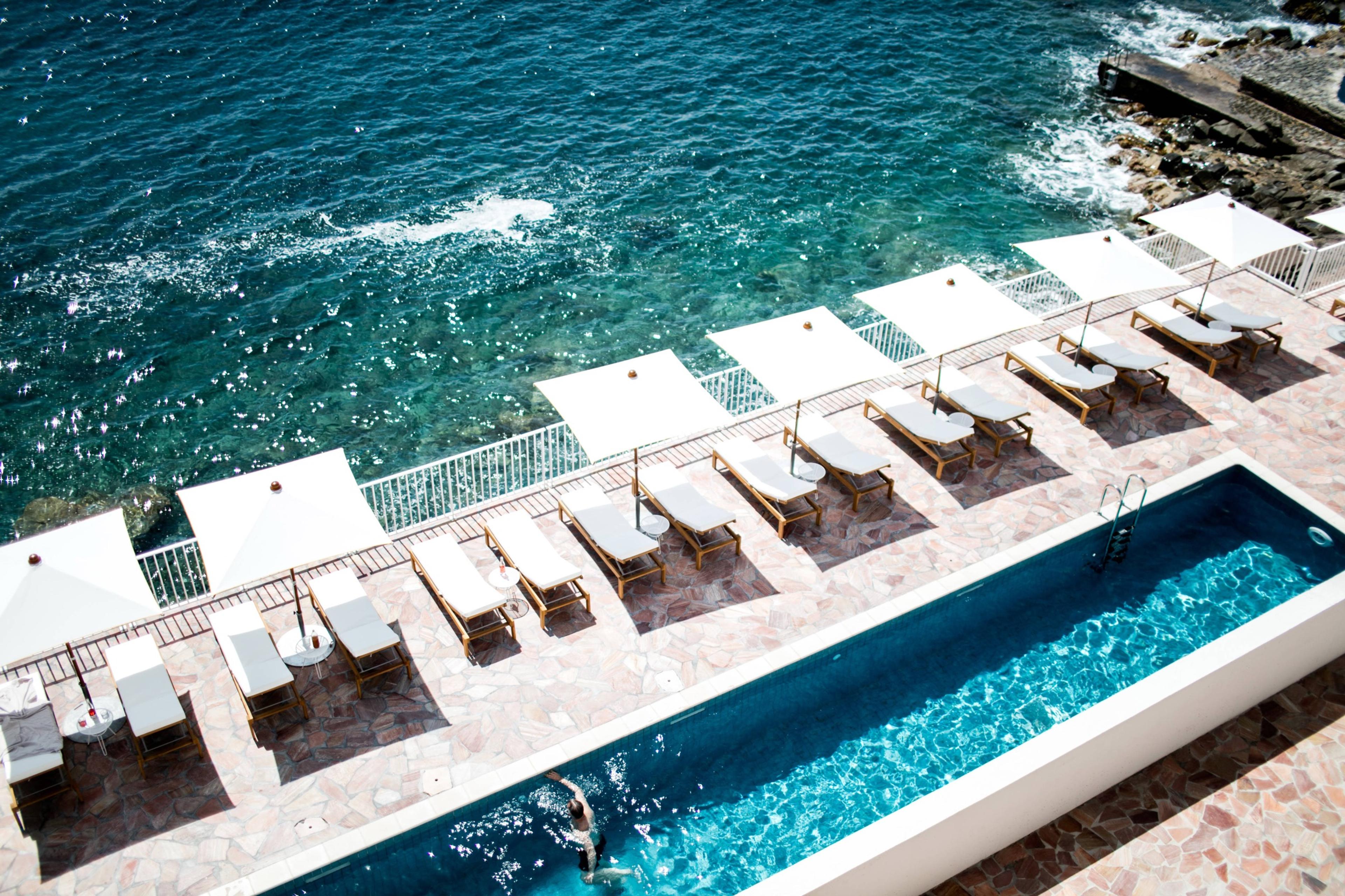 Looking down on a rectangular pool beside the sea, with rows of sun loungers under white parasols along the terrace edge, and turquoise water and rocks beyond.