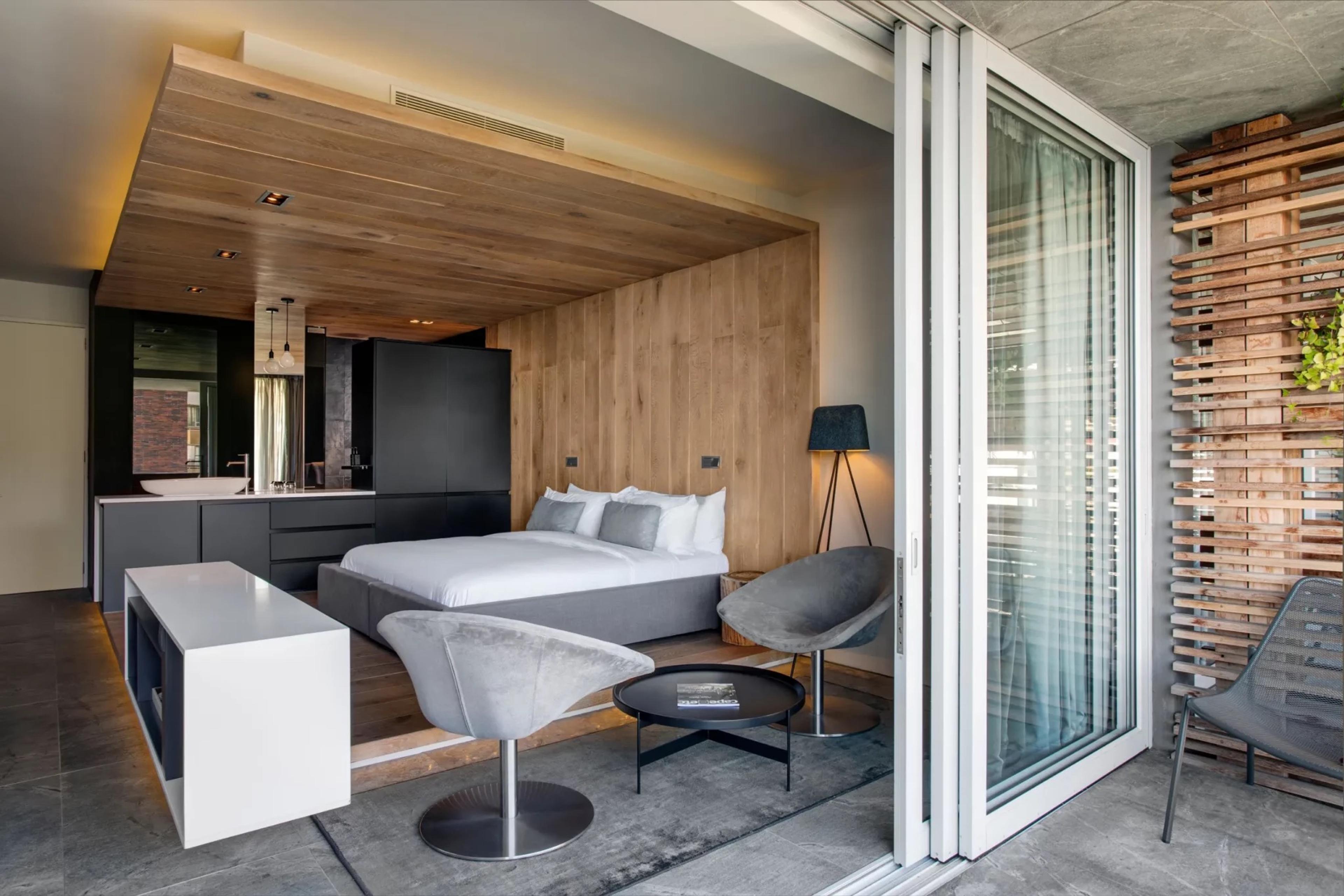 A hotel room with a king bed, a black kitchenette unit, grey swivel chairs and a glass wall sliding open to a screened balcony.