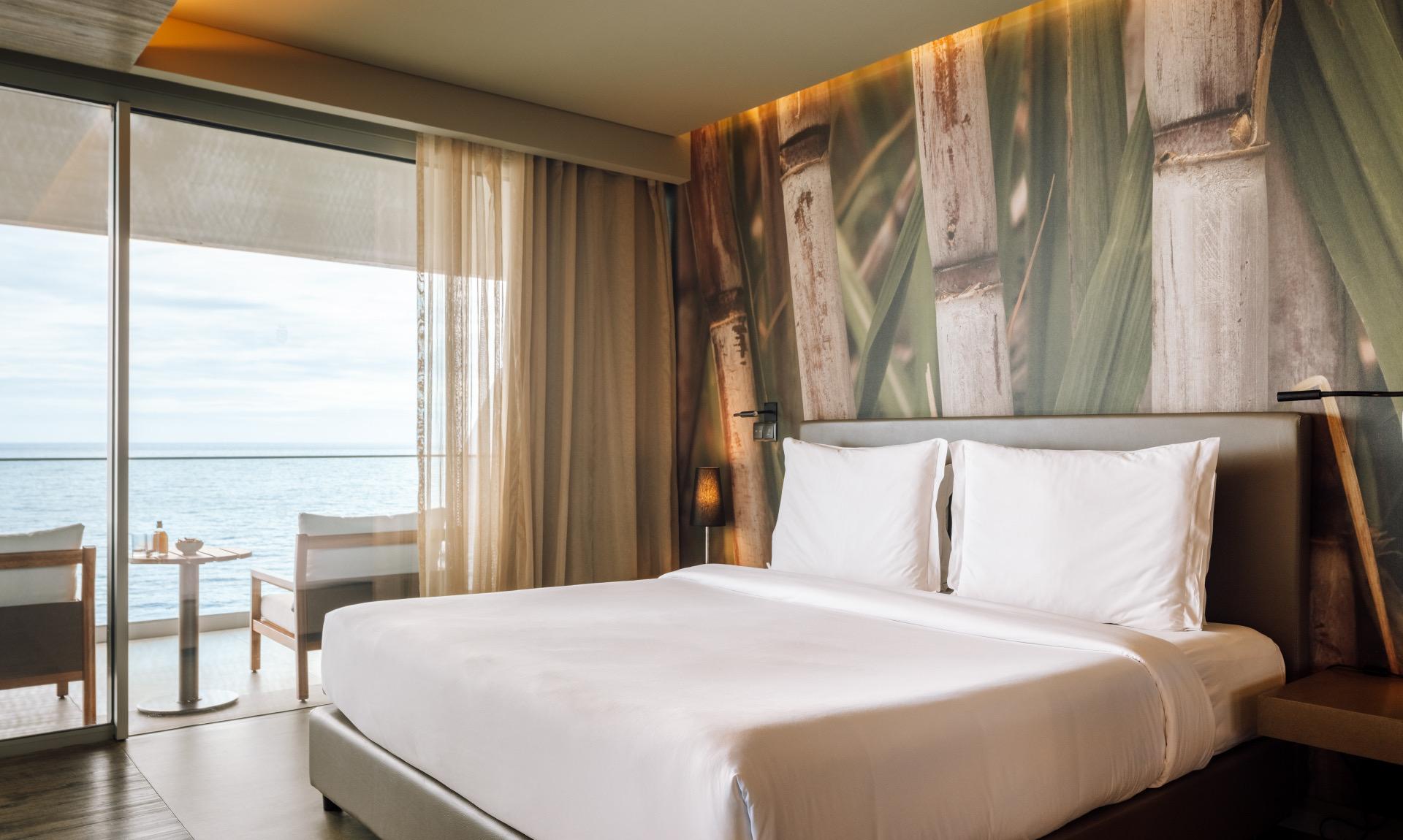 A bedroom with a bed set against a wall printed with a close up of sugar cane, and a sea view terrace beyond.
