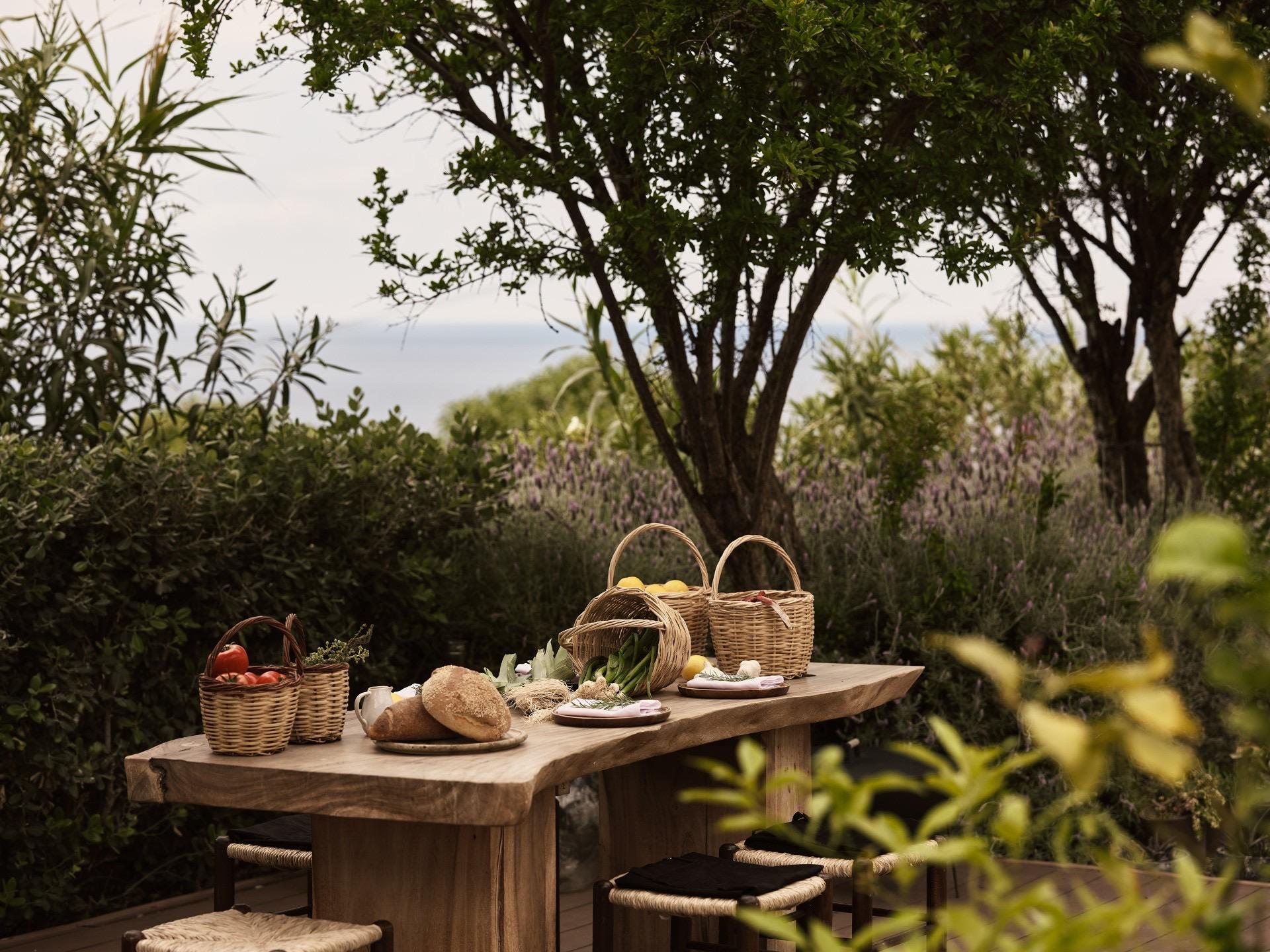 A rough wooden table in a garden holds baskets of tomatoes, lemons and vegetables, loaves of bread and a pitcher, with stools set around it.