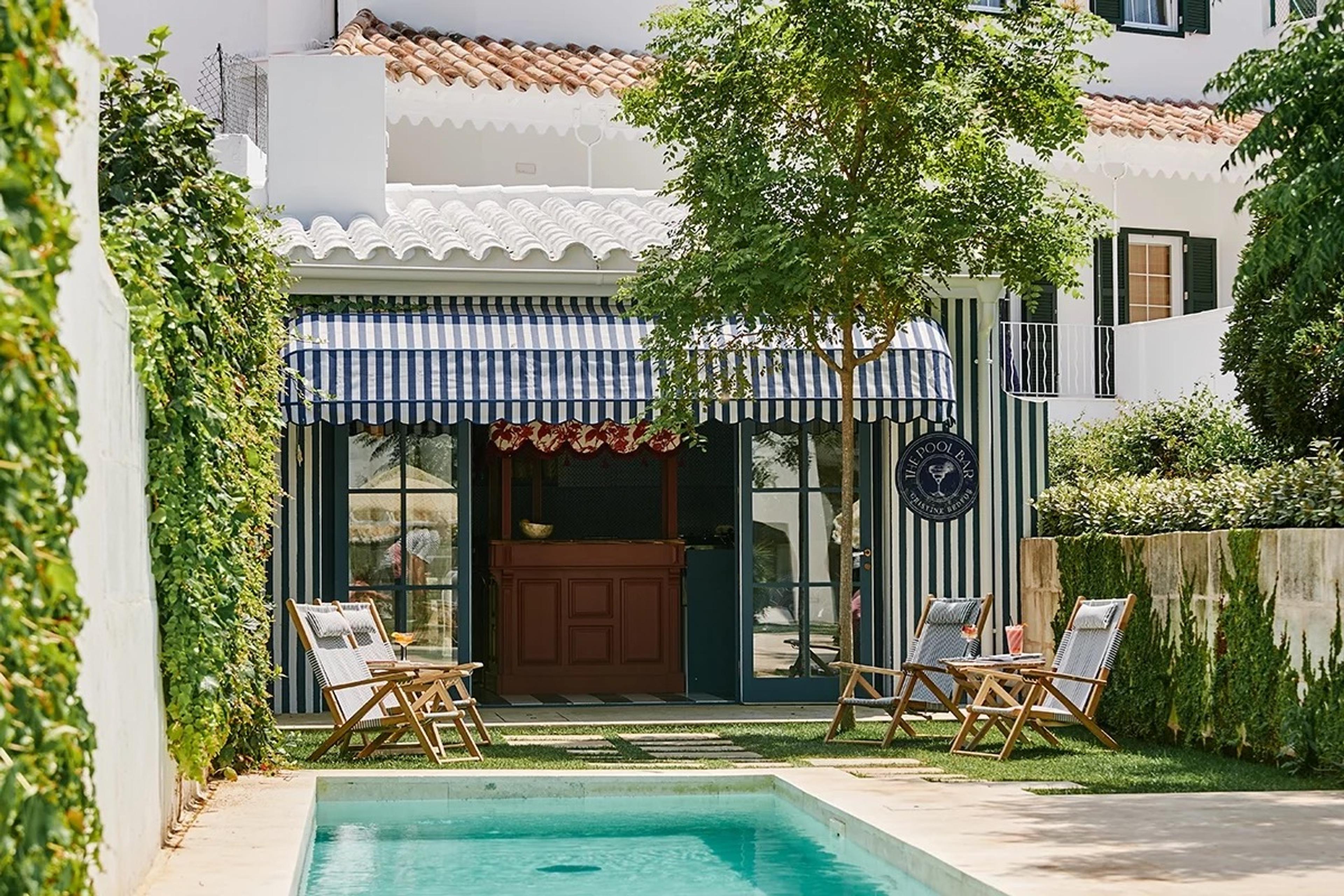 A tiled swimming pool below a stuccoed building with a blue and white striped awning over a pool bar, wooden deck chairs either side, and greenery climbing the courtyard walls.
