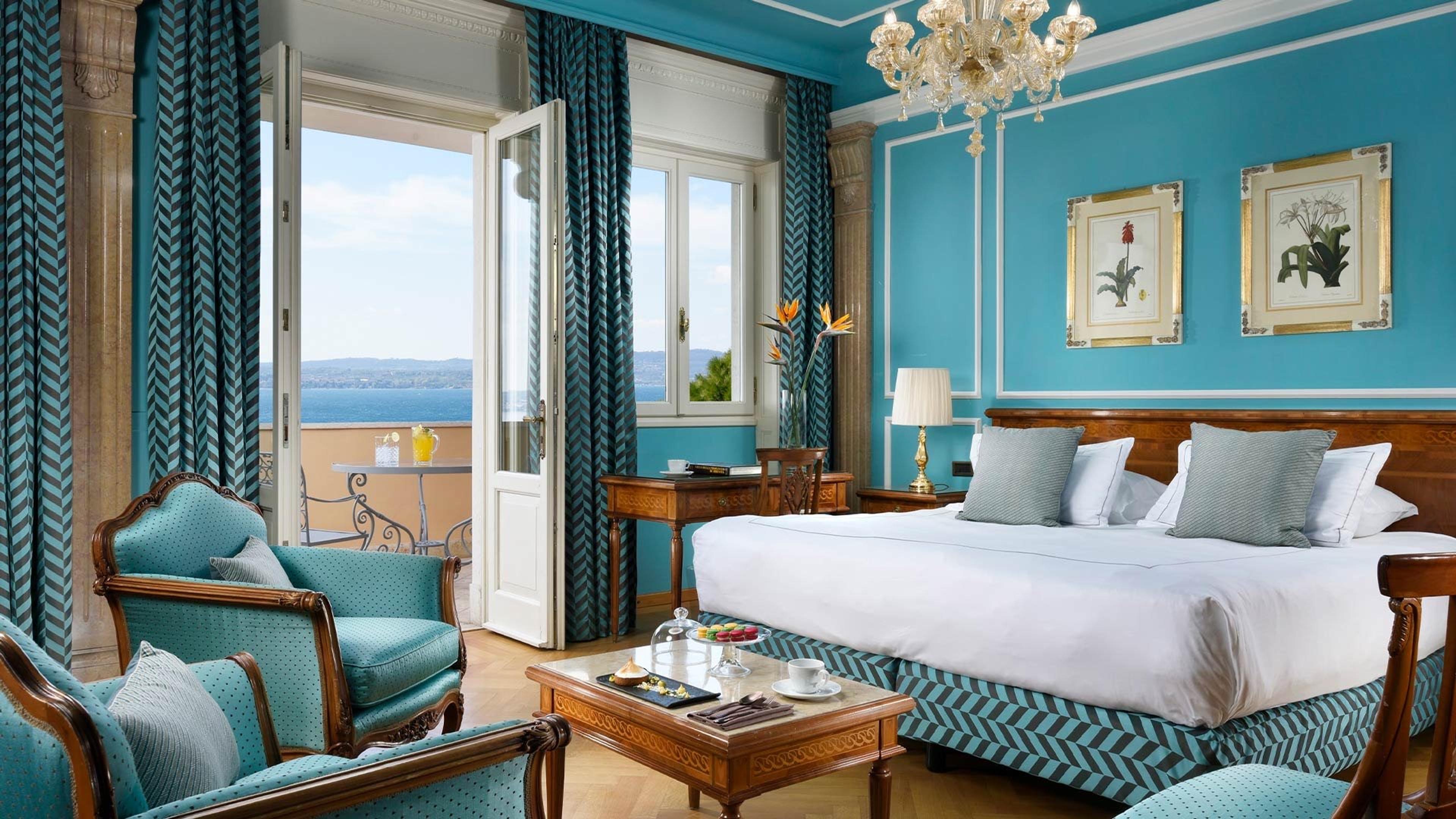 A teal-walled hotel suite with a canopy bed, armchairs, a crystal chandelier and a balcony overlooking a lake.