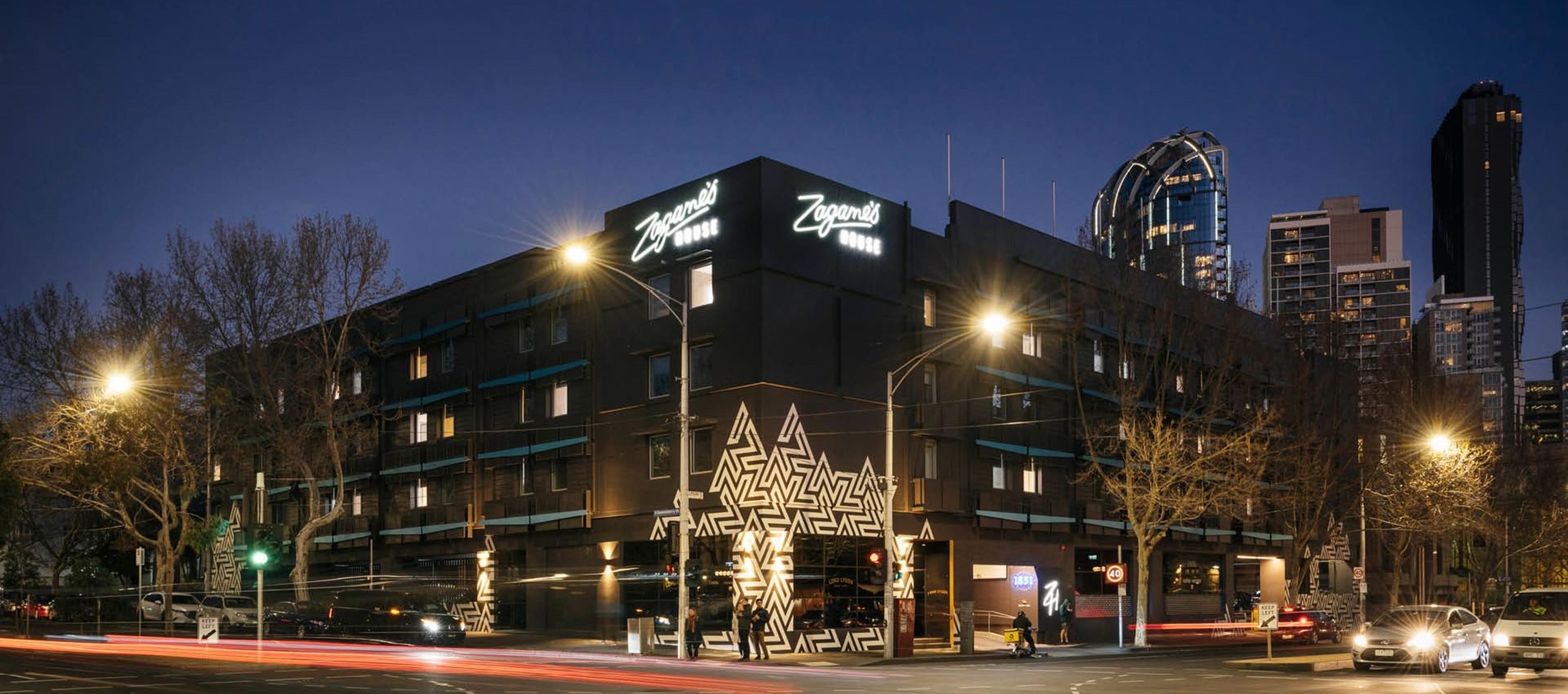 Zagame's House hotel building at night, lit signage above a street level entrance with a geometric mural, light trails from passing traffic