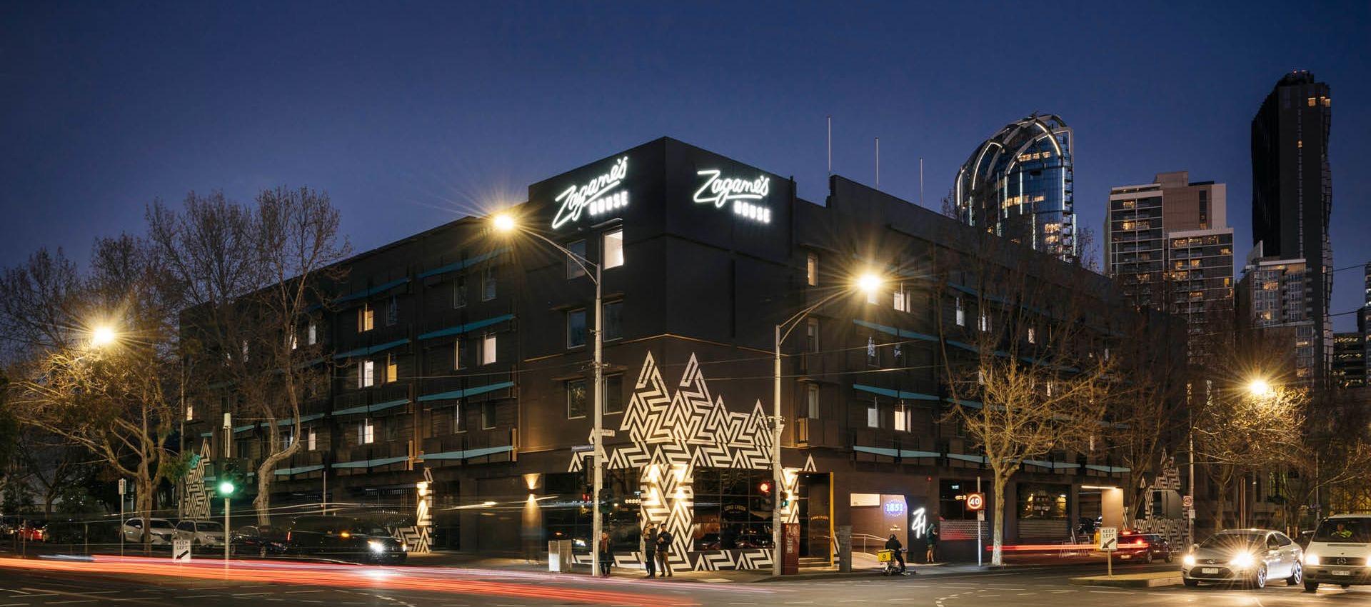 Zagame's House hotel building at night, lit signage above a street level entrance with a geometric mural, light trails from passing traffic