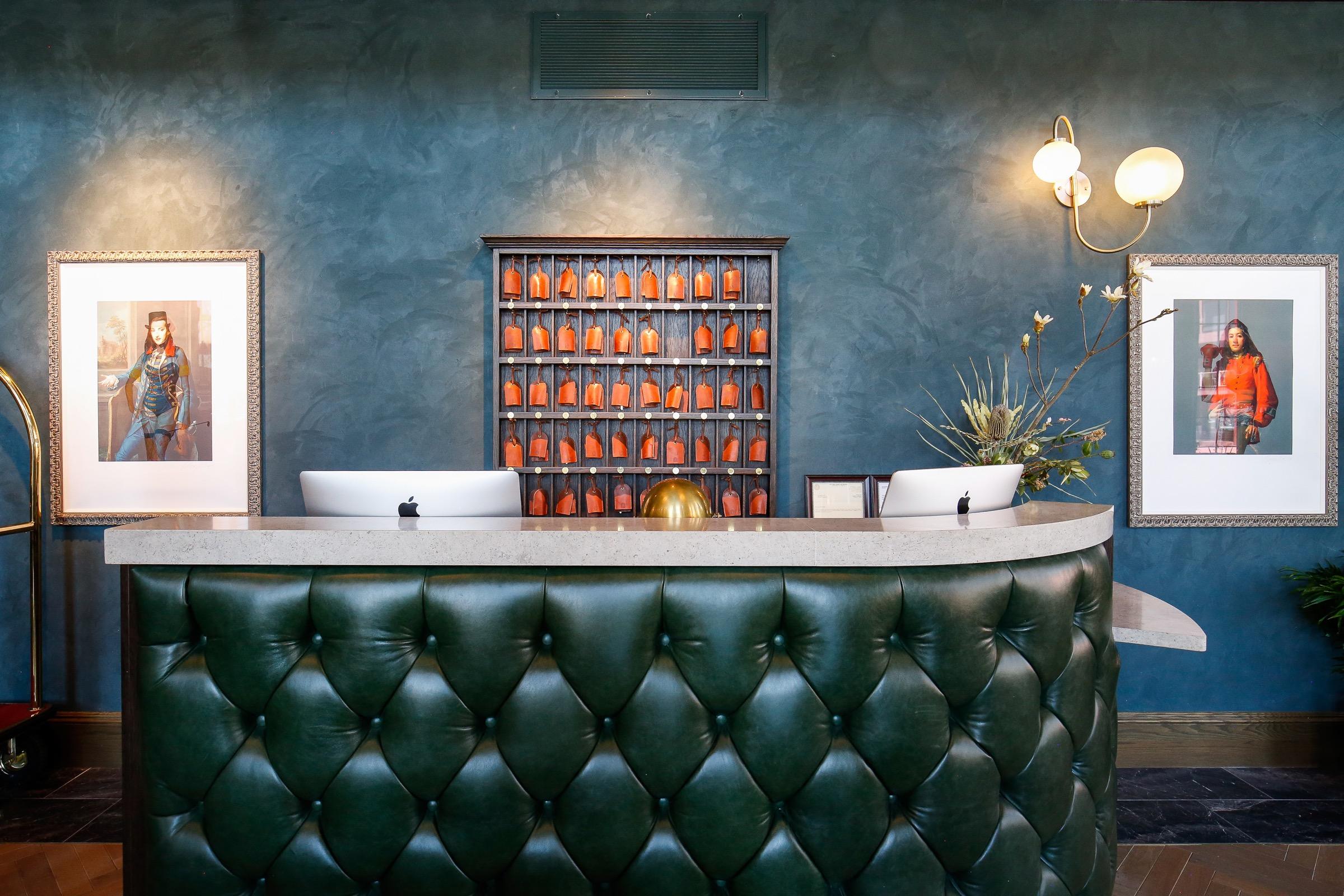 A tufted leather reception desk below a wall of numbered key cubbies, flanked by two framed illustrated portraits.
