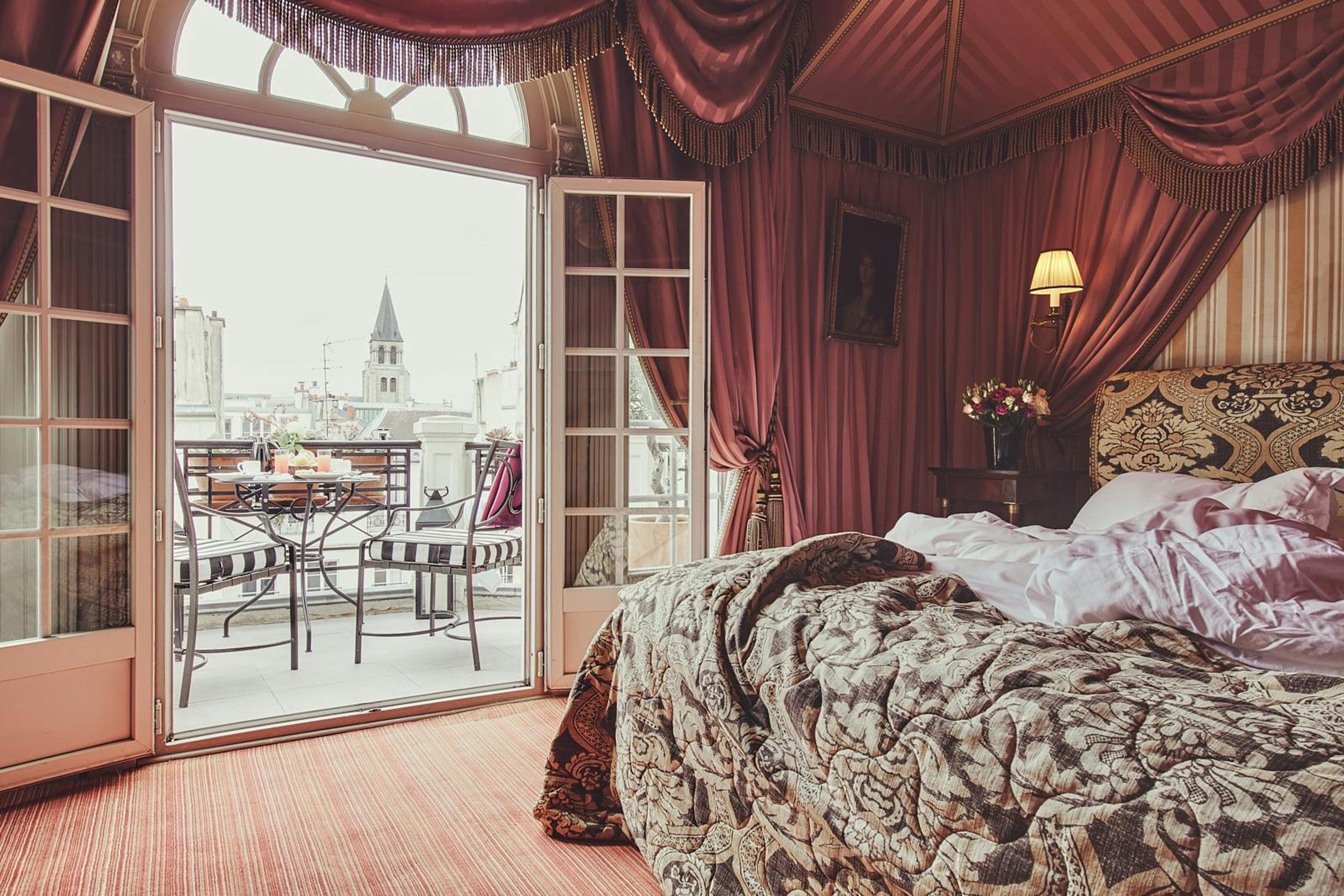 A bedroom with ornate drapery and a patterned bedspread, its French doors open onto a small balcony overlooking rooftops and a church spire.