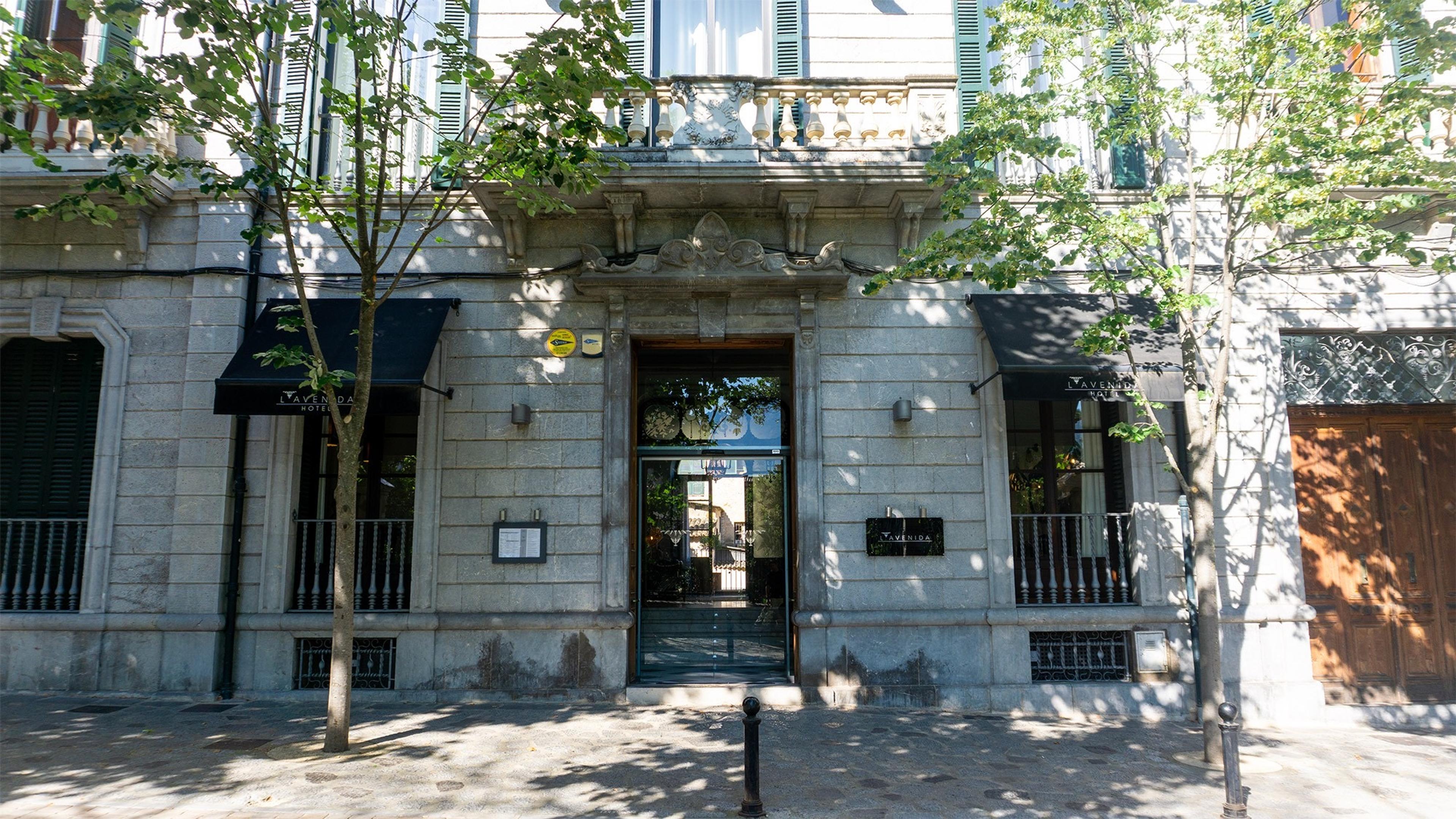 The stone corner facade of the hotel, with arched windows, iron balconies, a columned entrance, and small trees shading the pavement.