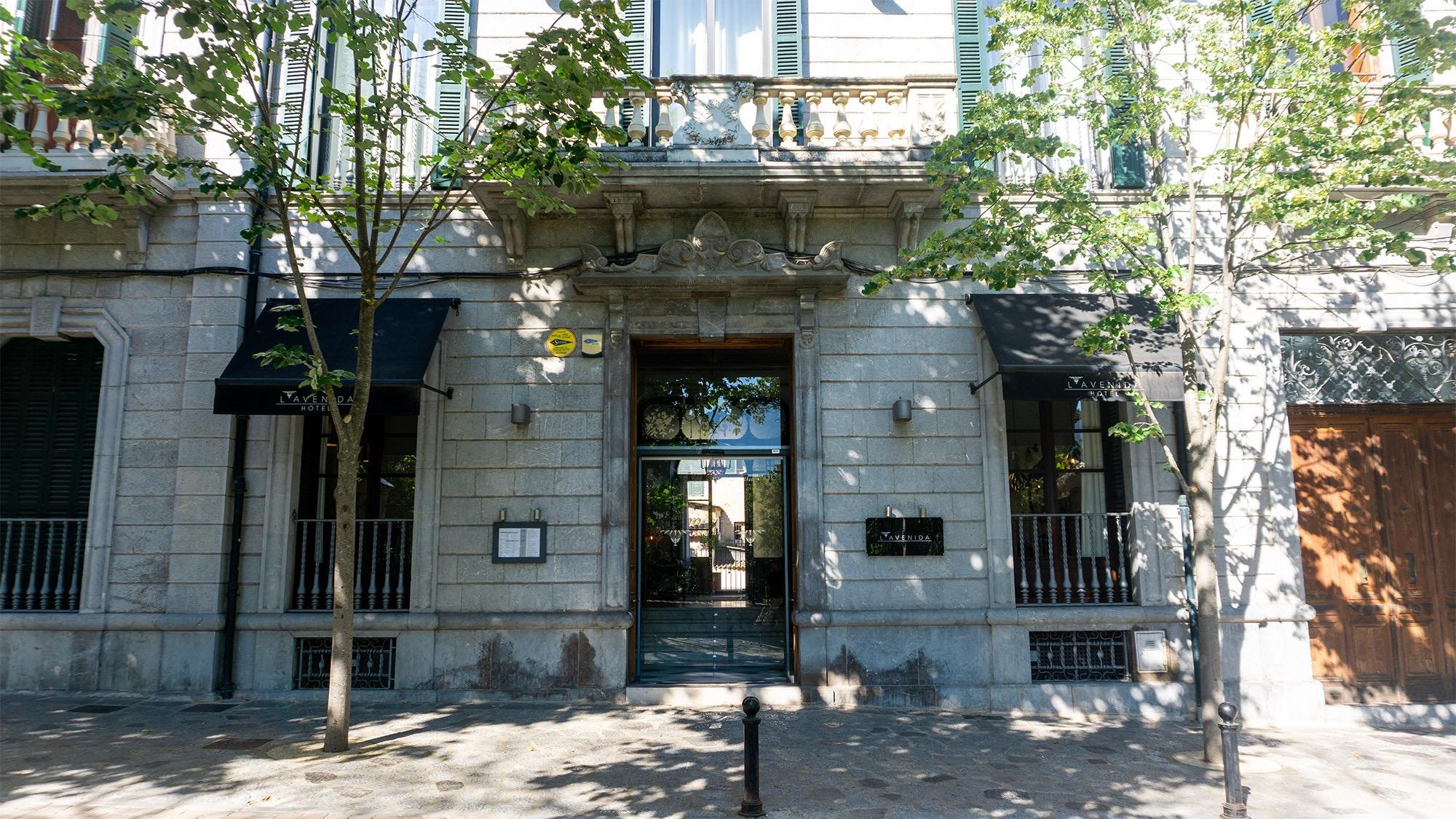 The stone corner facade of the hotel, with arched windows, iron balconies, a columned entrance, and small trees shading the pavement.