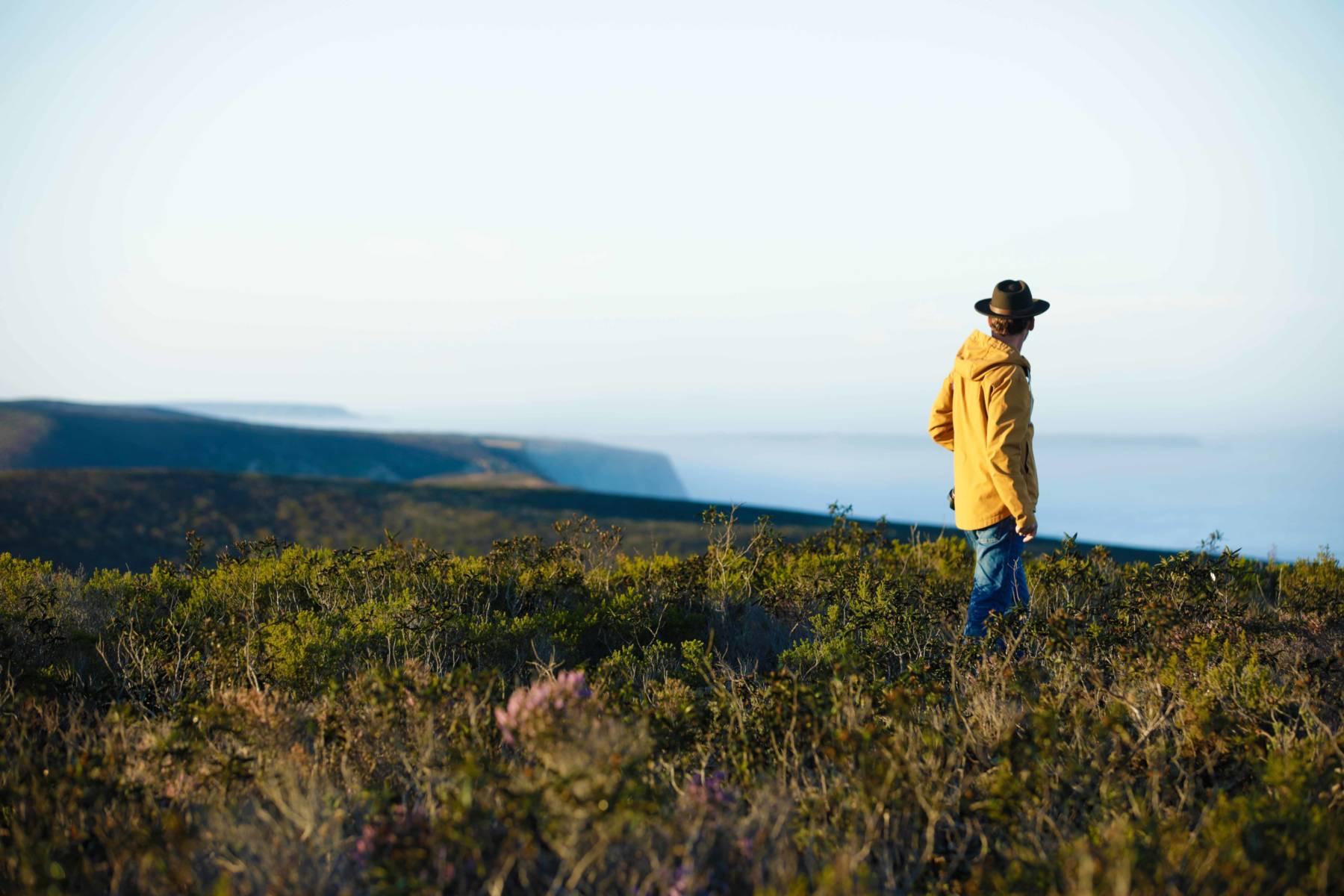 A man in a yellow jacket and hat walking through coastal scrubland, looking out toward cliffs and the sea.