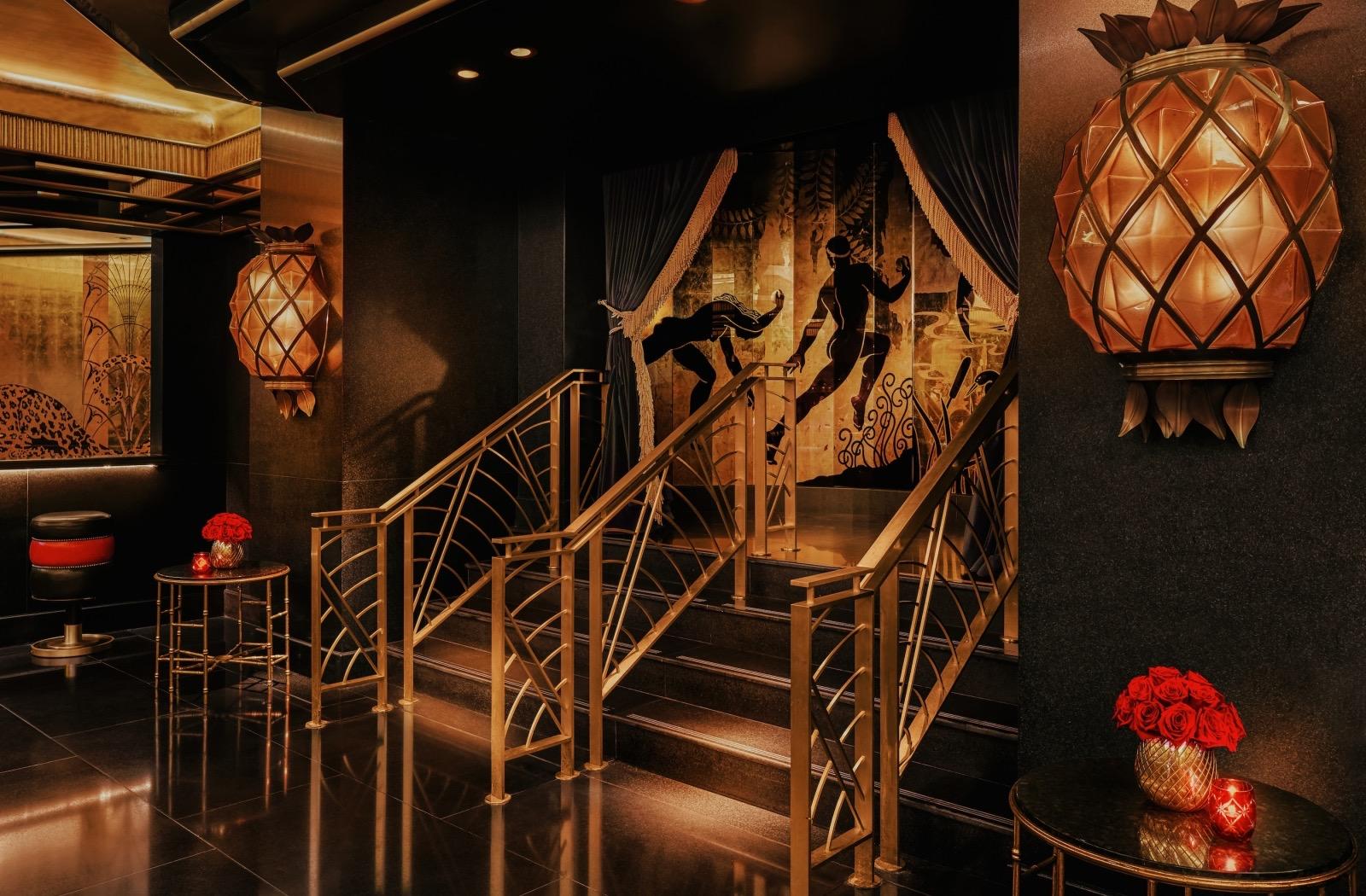 A dark lobby with a gold-railed staircase, pineapple-shaped wall lights and a painted mural of figures, with red roses on a side table.