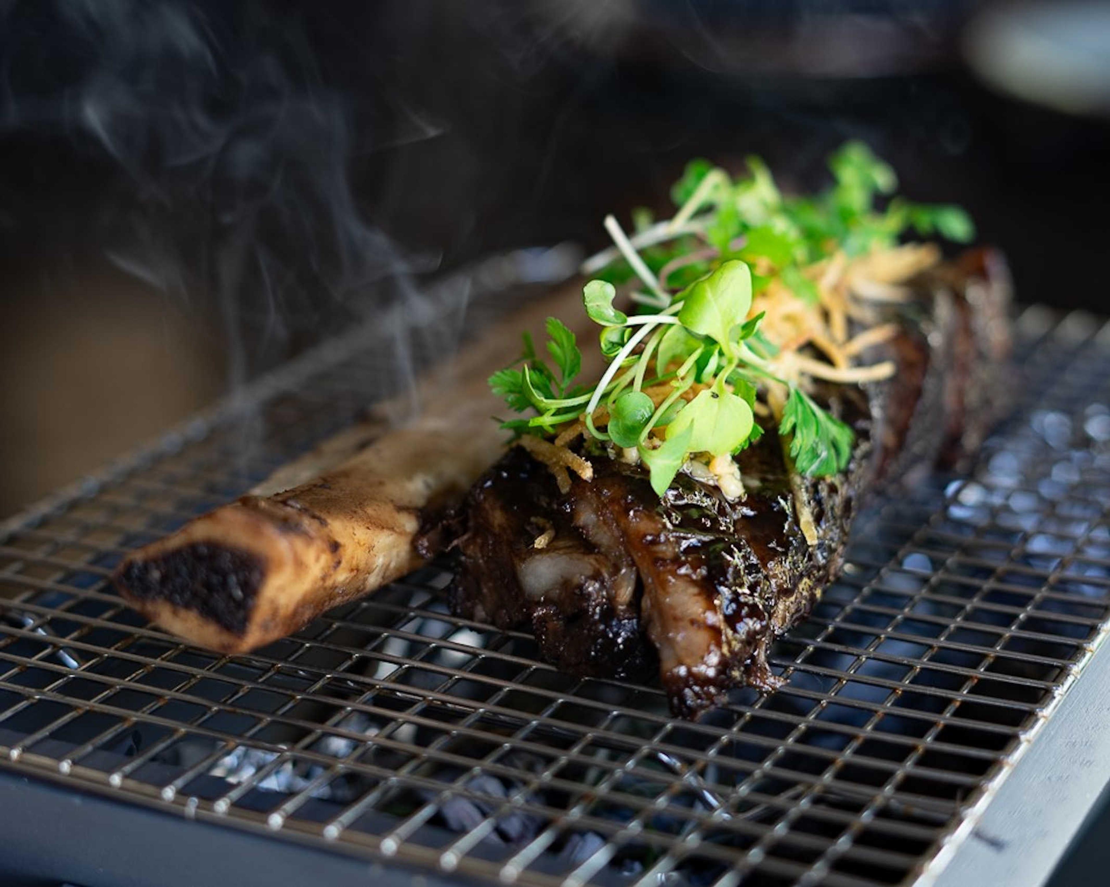 A charred bone-in short rib on a charcoal grill, topped with fresh herbs and crispy shallots, with smoke rising around it.