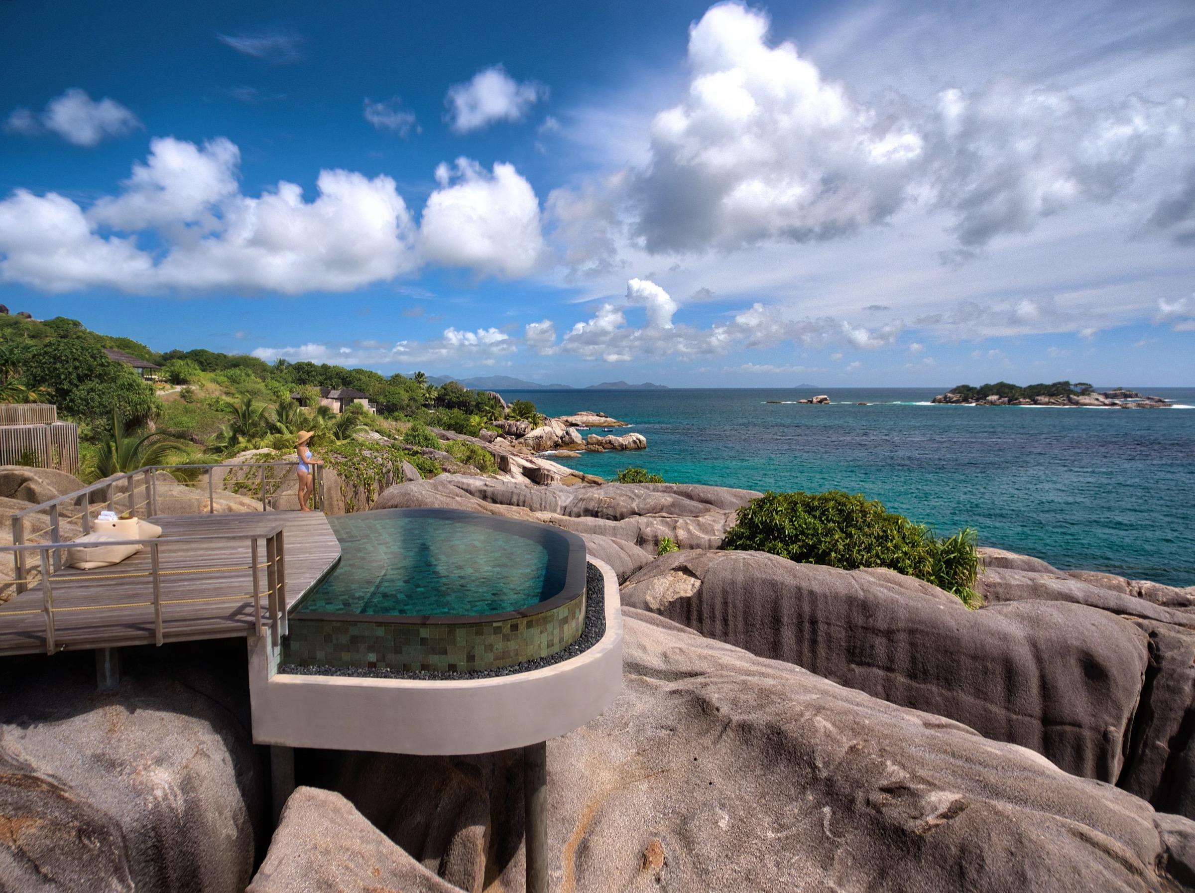 A wooden-decked pool overlooking a rugged granite shoreline, clear blue-green sea and small islands.