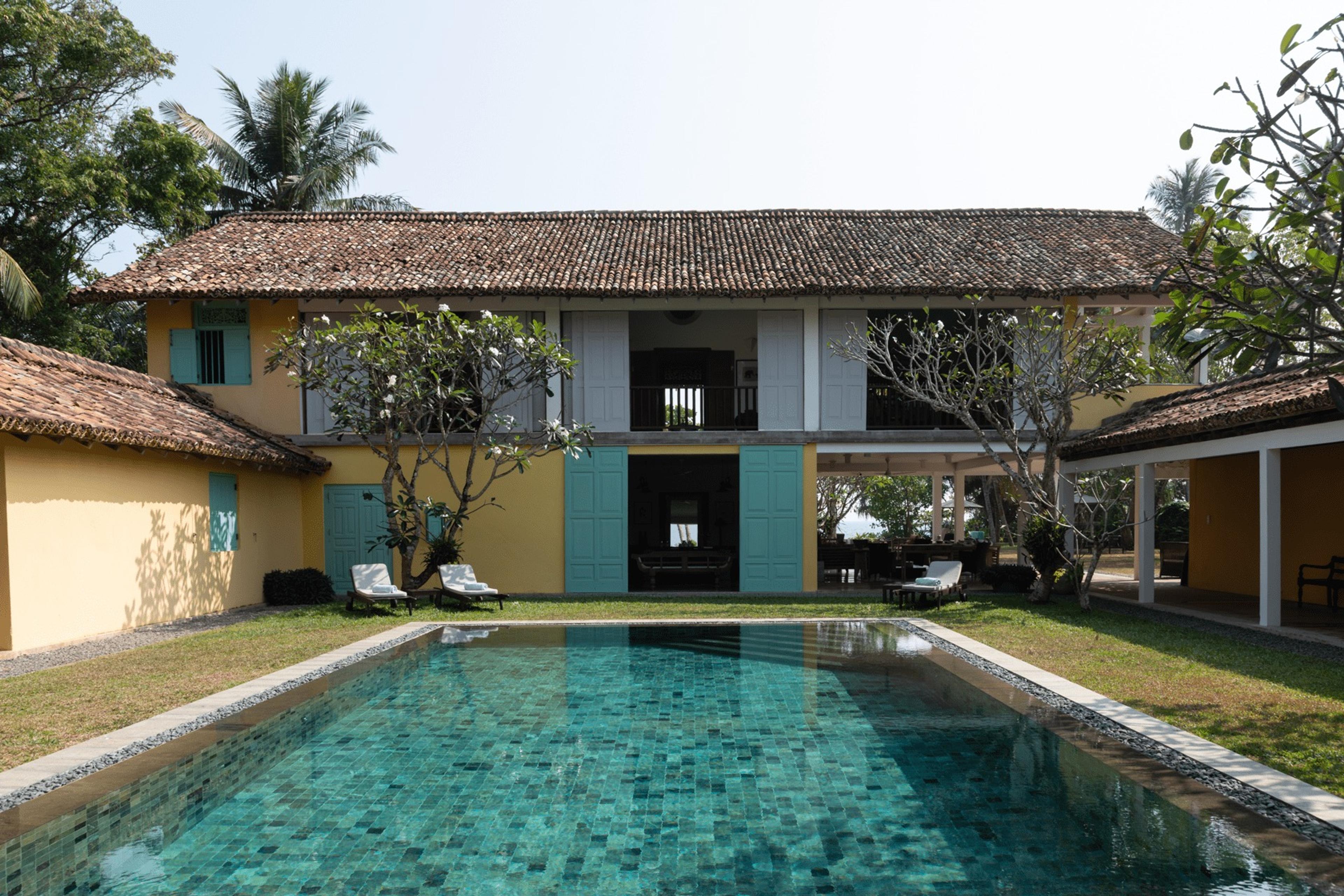 A two storey yellow house with teal shuttered doors and a terracotta tiled roof stands behind a swimming pool, with rattan sun loungers and palm trees around it.