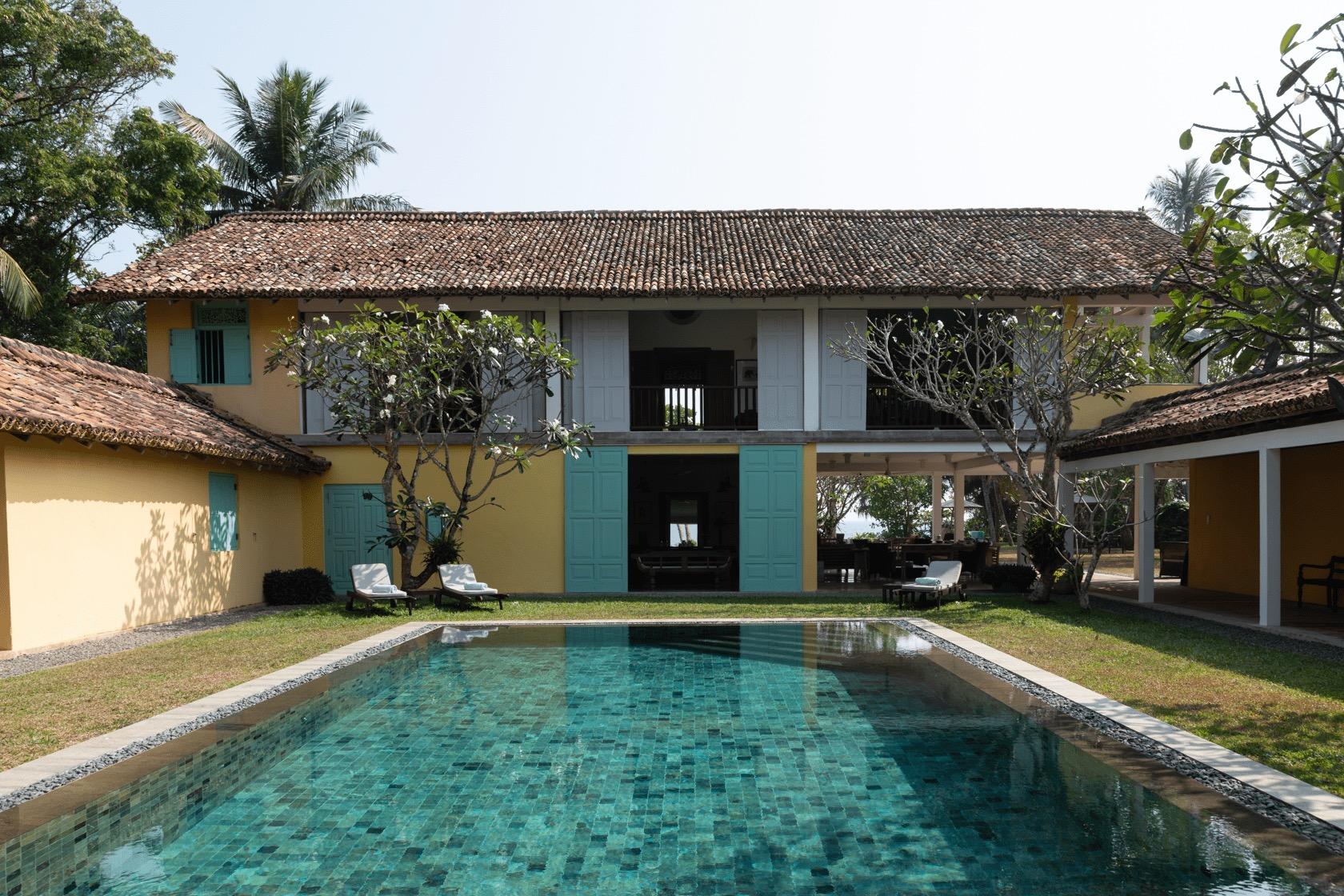A two storey yellow house with teal shuttered doors and a terracotta tiled roof stands behind a swimming pool, with rattan sun loungers and palm trees around it.