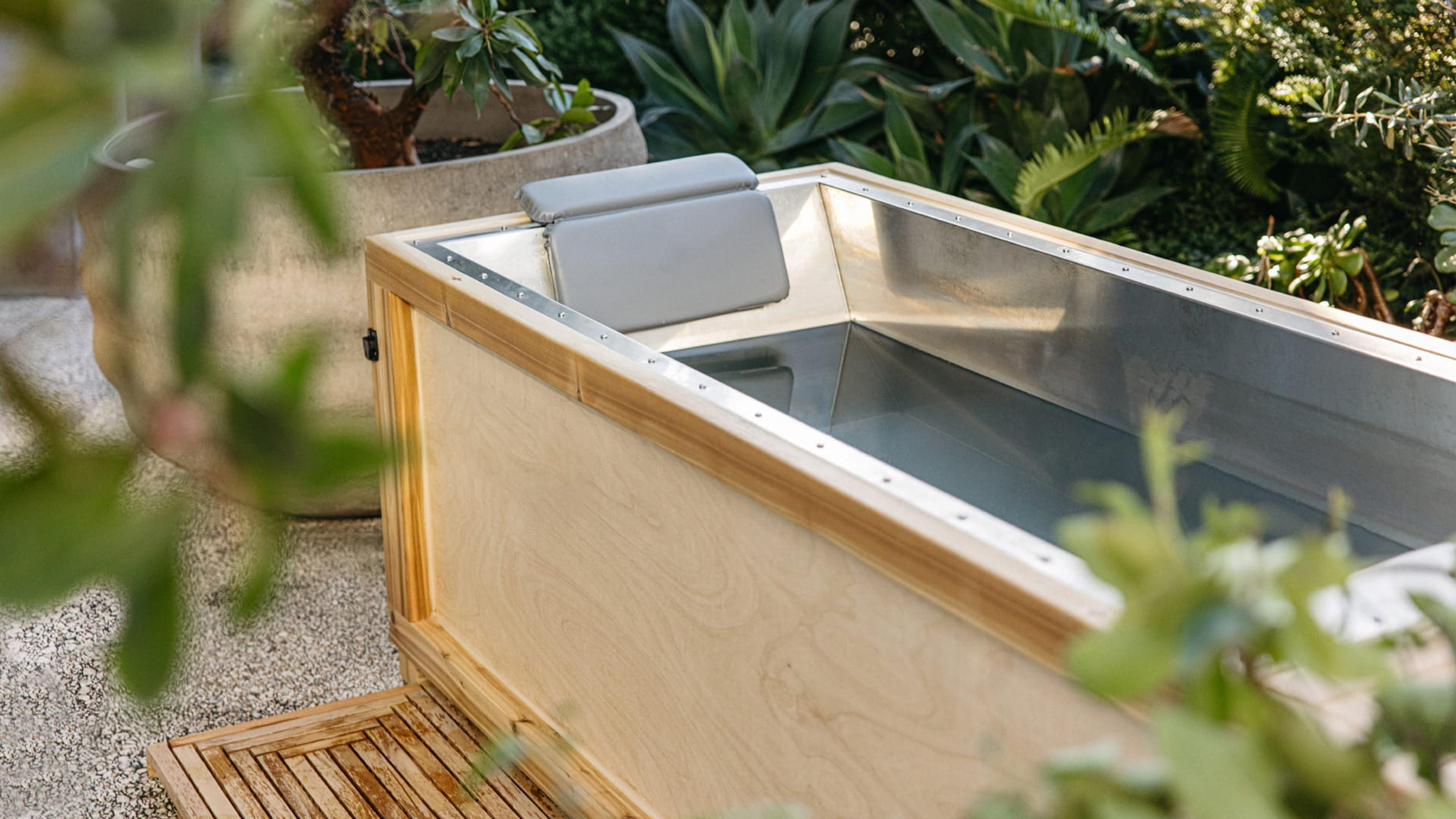 An outdoor wooden cold plunge tub with a metal lining, set on a stone deck among potted palms and agave.