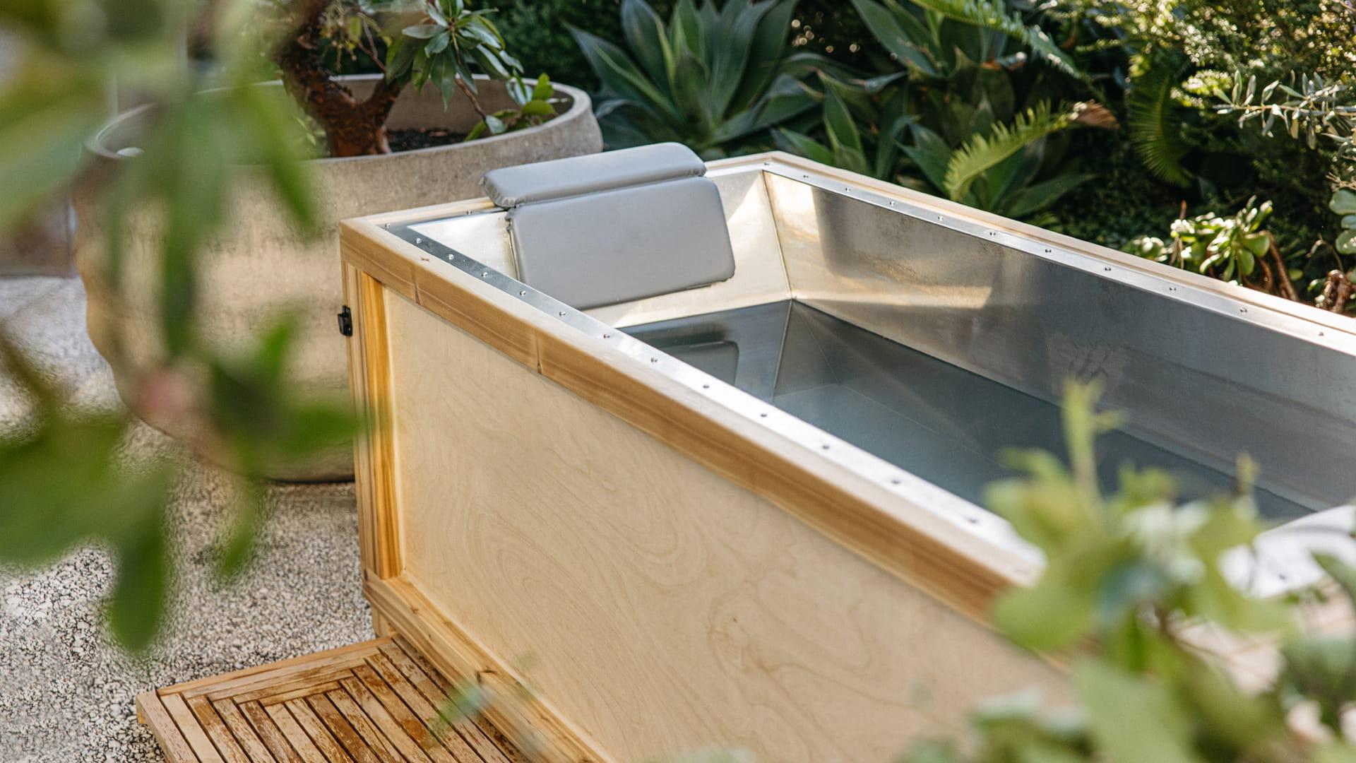 An outdoor wooden cold plunge tub with a metal lining, set on a stone deck among potted palms and agave.