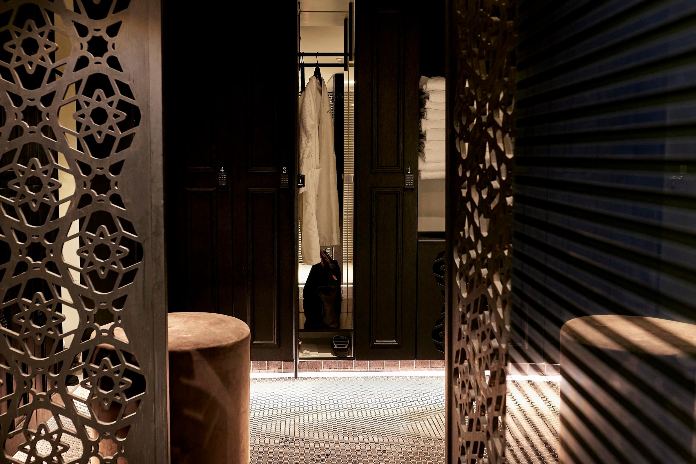 Two dark wooden locker doors stand open between patterned metal screens, showing a hanging robe, folded towels and a bag inside, with stools on the tiled floor.