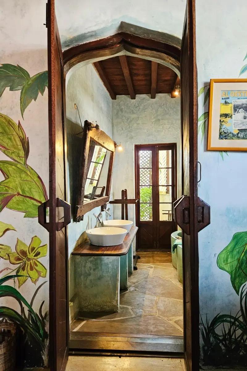 two open wooden doors painted with botanical murals, leading to a bathroom with twin vessel sinks, a tilted mirror and French doors to a balcony