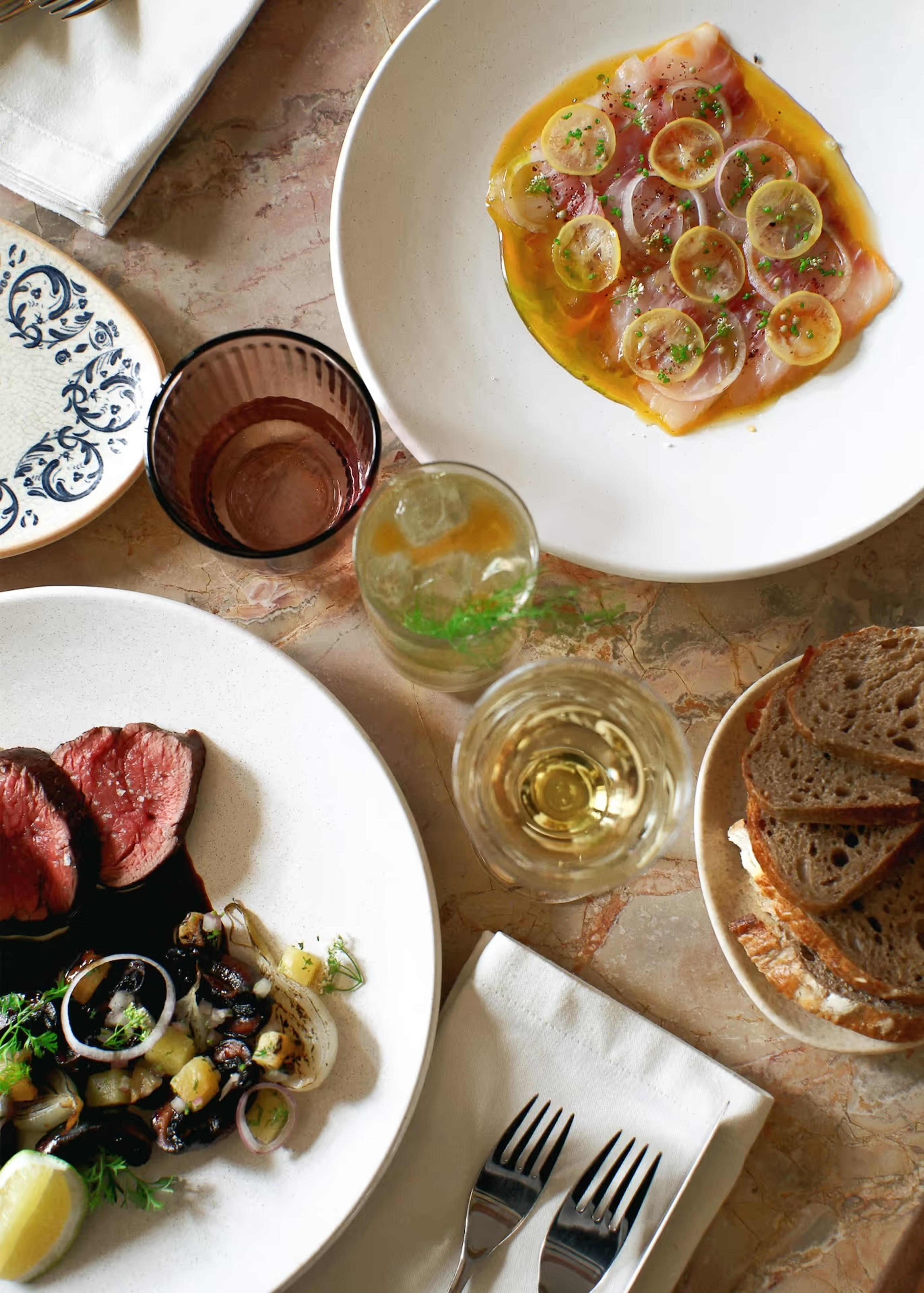 Shared plates of sliced beef, seafood and bread on a marble table with glasses of wine