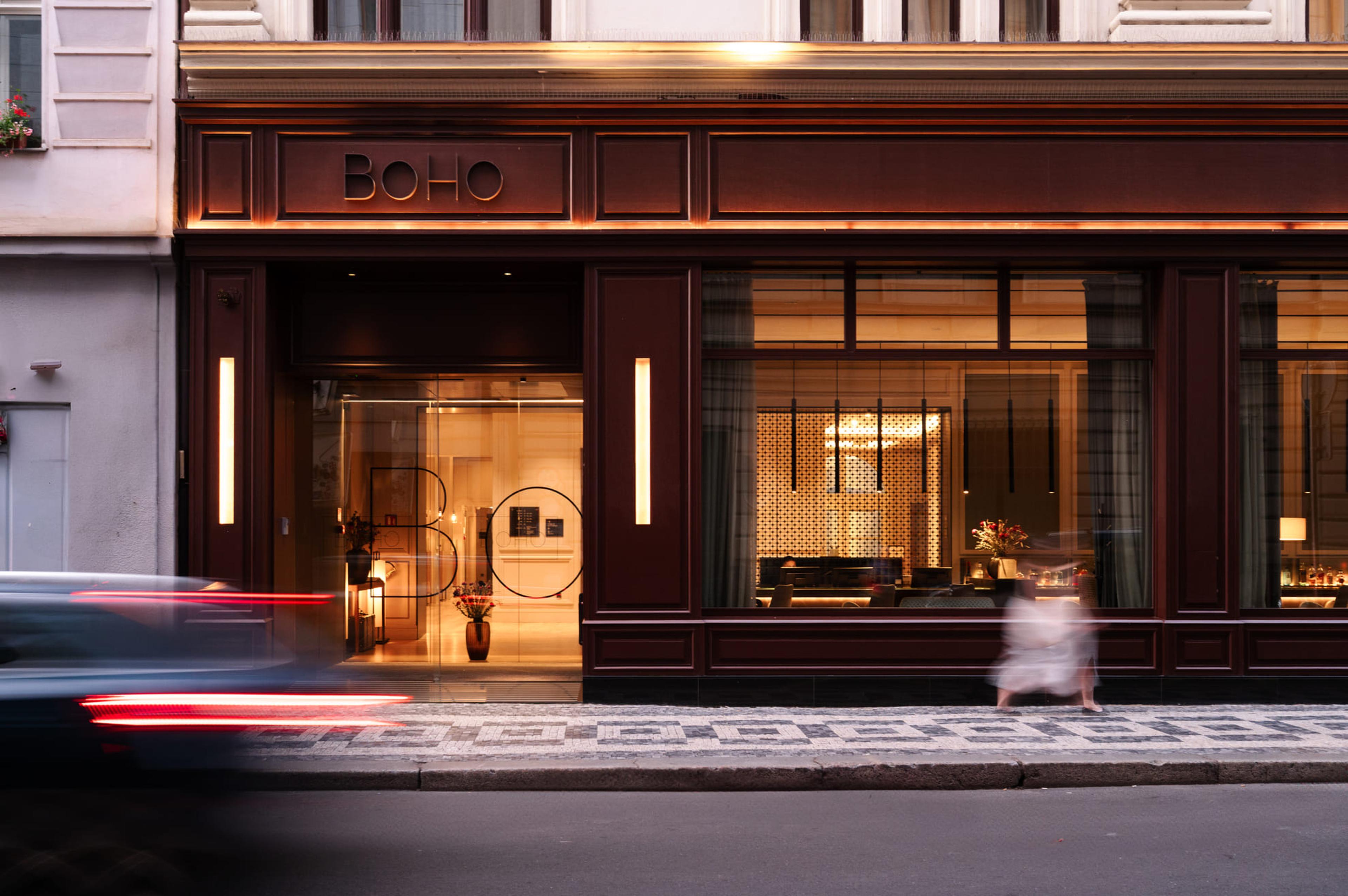 The front of the building at dusk, with the name BOHO on a dark brown shopfront, a lit doorway on the left, and a blurred car passing in the foreground.