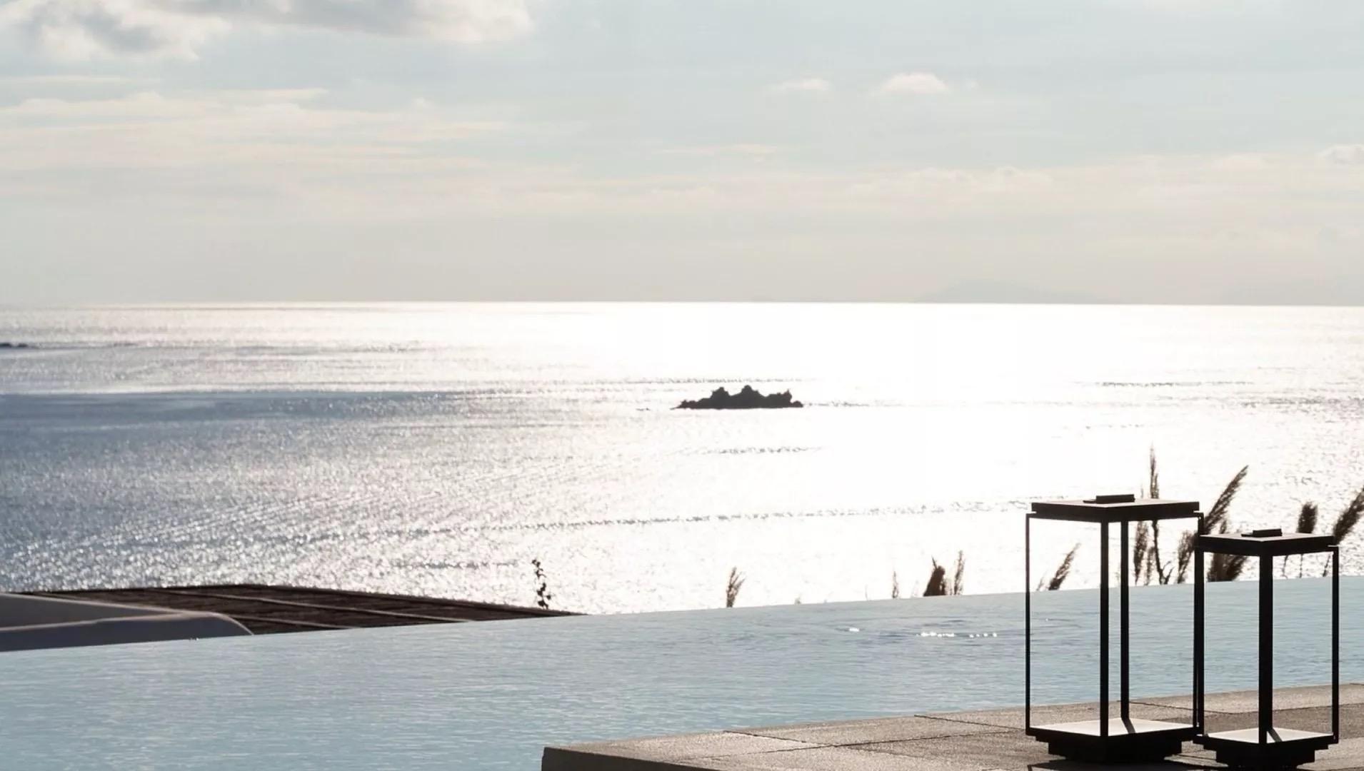 The sea at sunset seen past a pool's edge and two dark lanterns, with a small rocky islet on the horizon.
