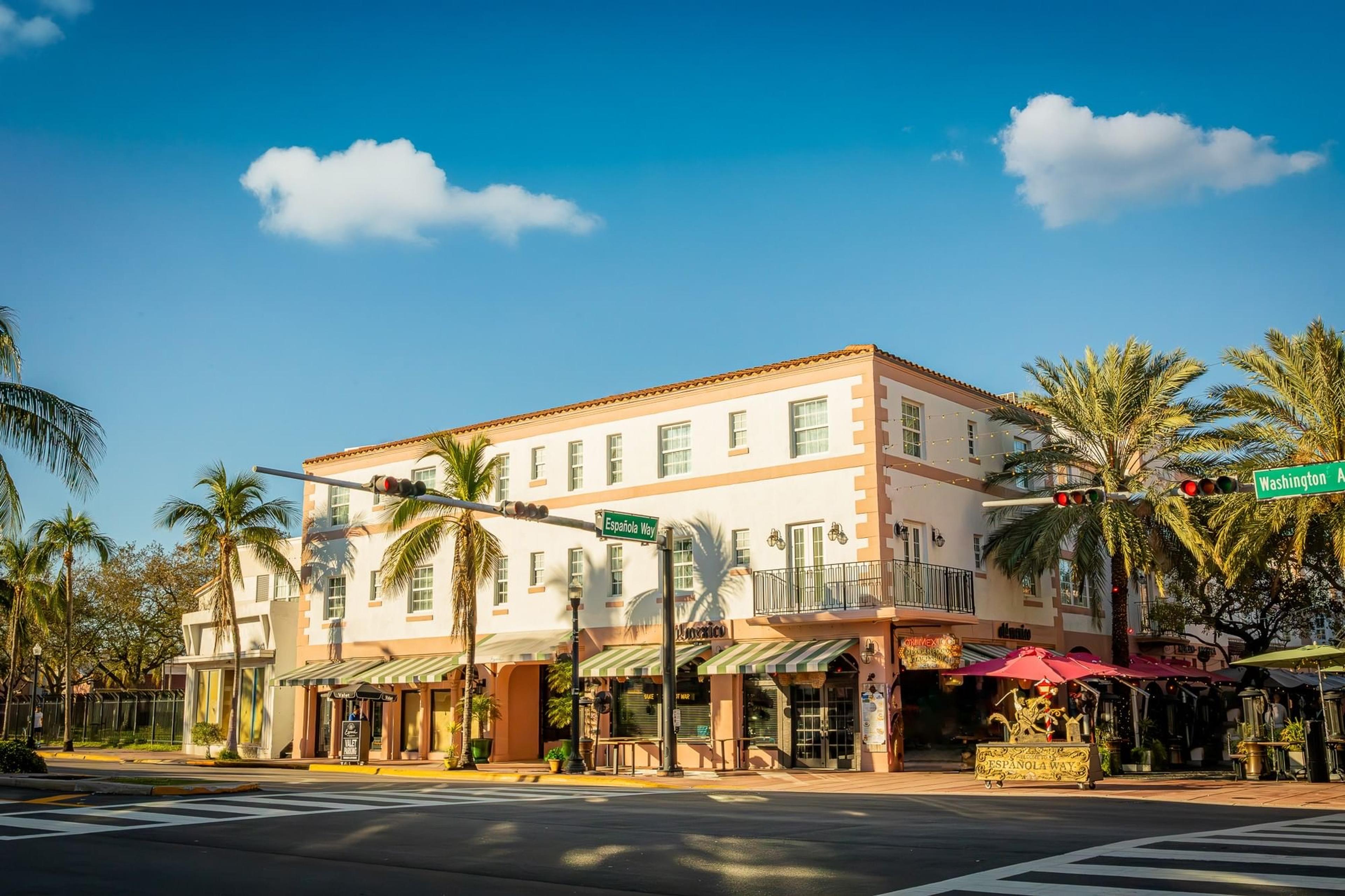 Looking up at a three-storey cream and coral hotel building on a sunny street corner, with palm trees, striped awnings, and street signs for Espanola Way and Washington Avenue.