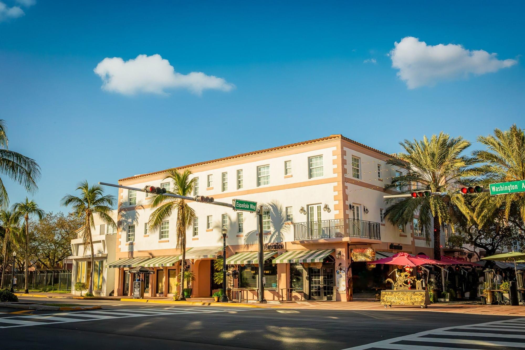 Looking up at a three-storey cream and coral hotel building on a sunny street corner, with palm trees, striped awnings, and street signs for Espanola Way and Washington Avenue.