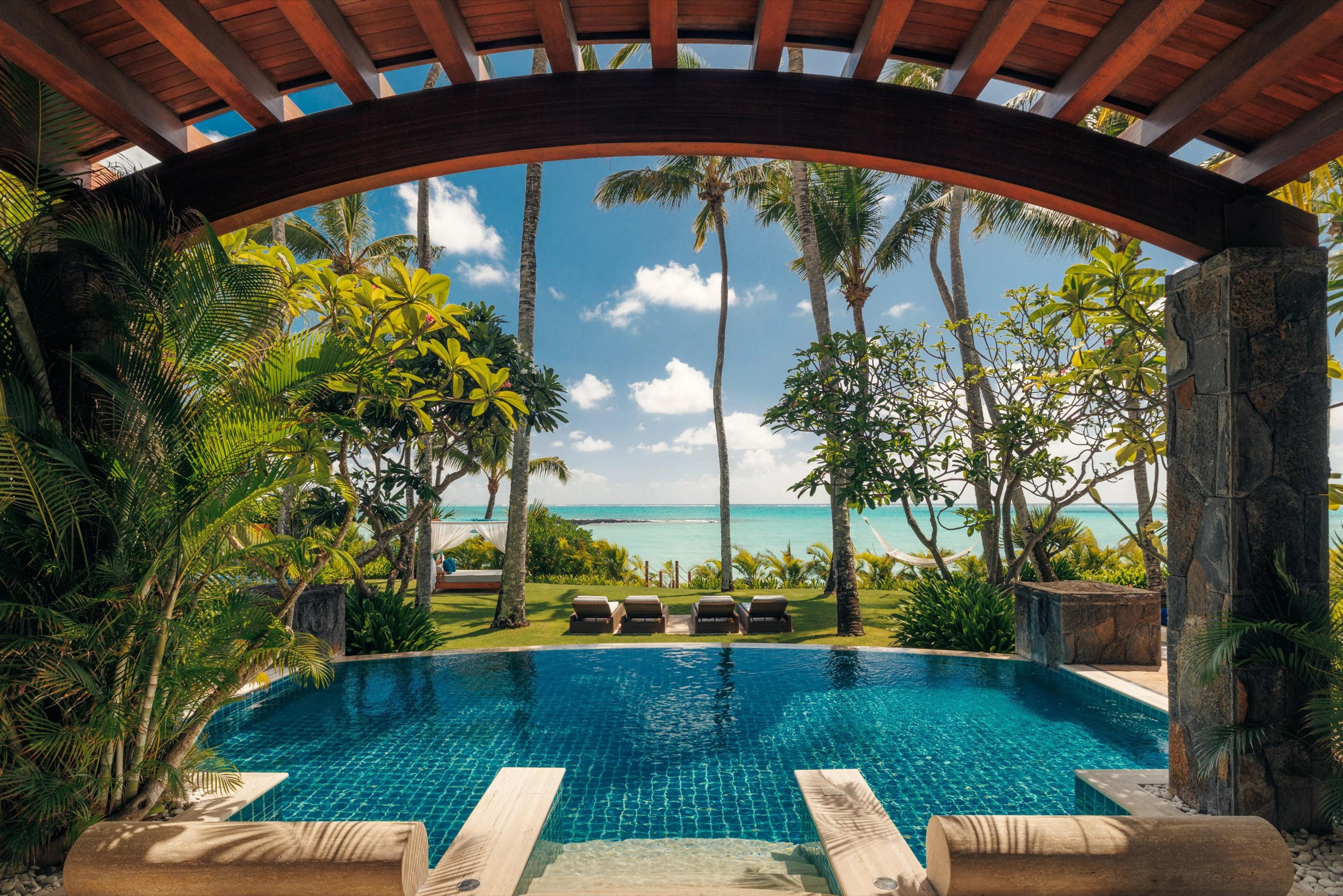 A wooden pergola frames an infinity pool and sun loungers leading to palm trees and the open sea.