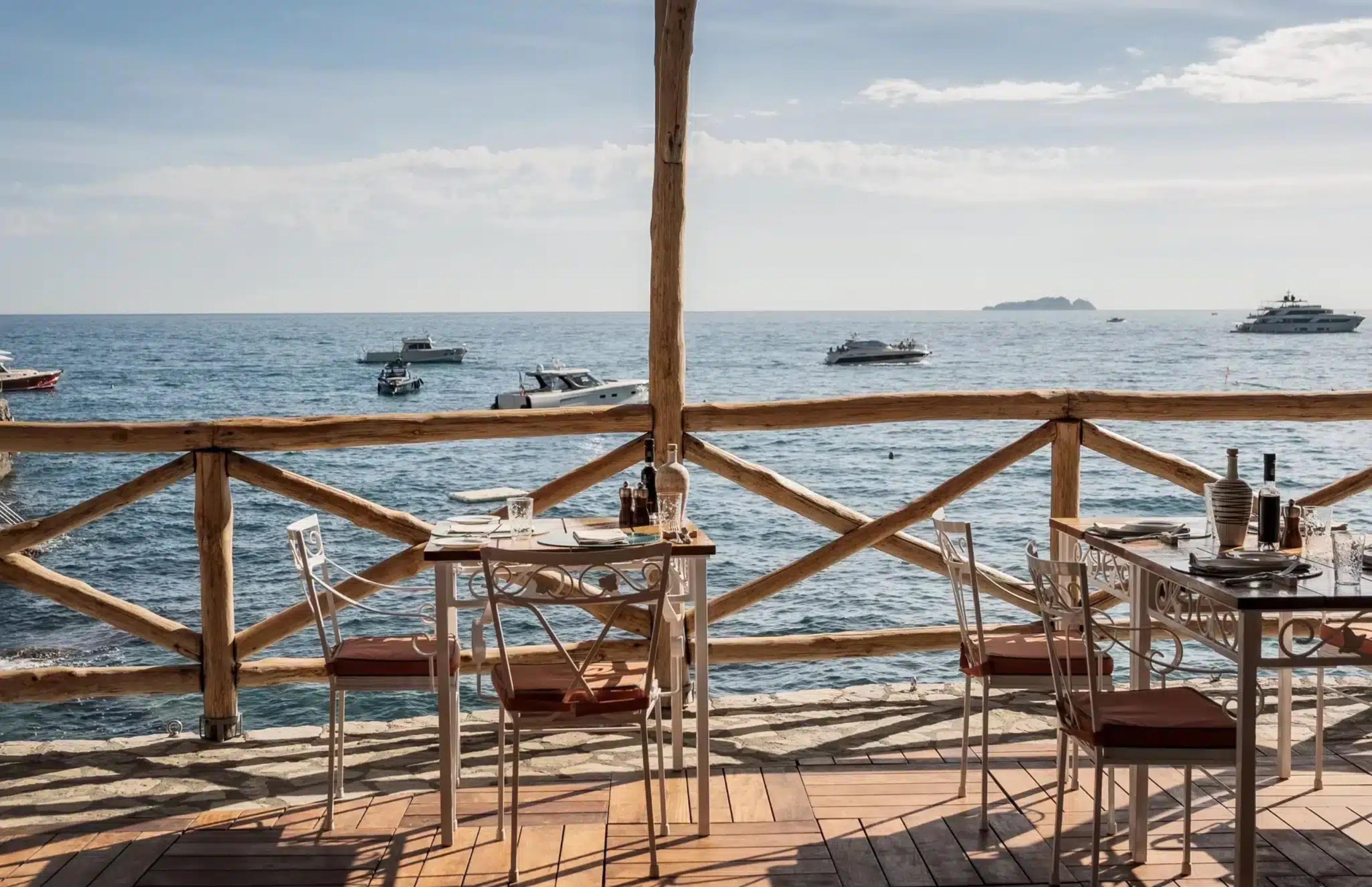 An empty restaurant terrace with wooden tables and chairs built over the sea, boats anchored in the water below.