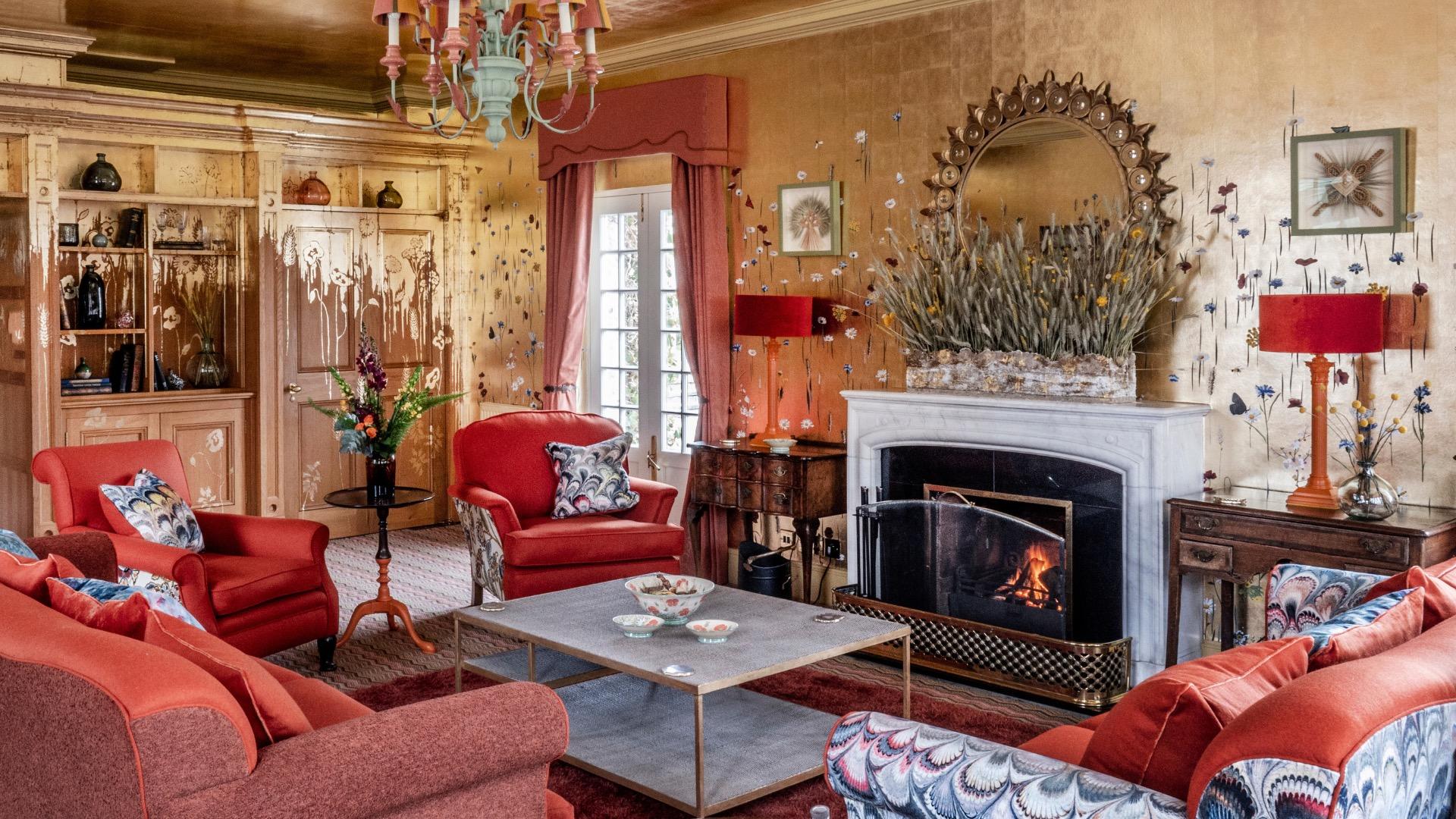 A living room with red armchairs and sofas around a low table, facing a white marble fireplace with a lit fire and a round mirror above it