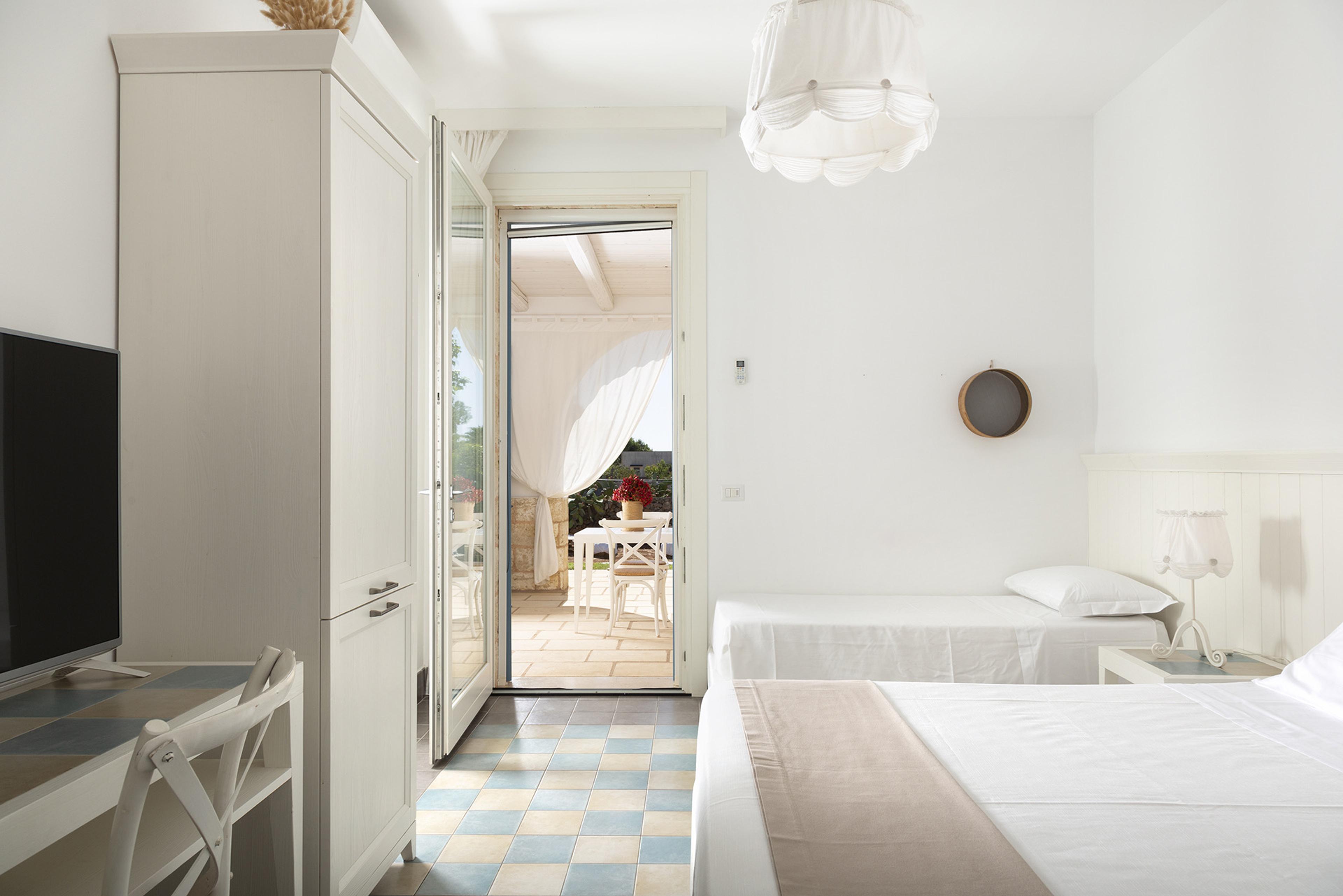 A bedroom with twin beds, a wardrobe and a television, its door open onto a veranda with a table set outside.
