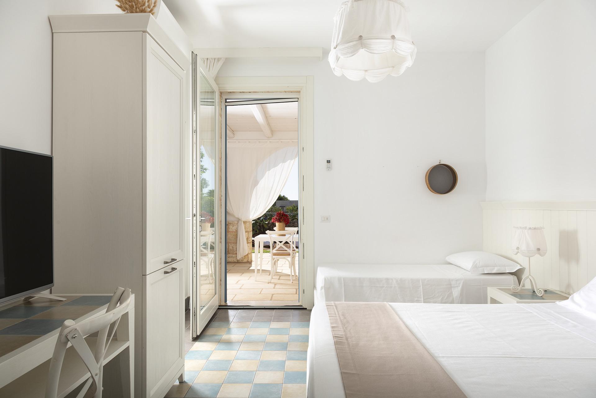 A bedroom with twin beds, a wardrobe and a television, its door open onto a veranda with a table set outside.