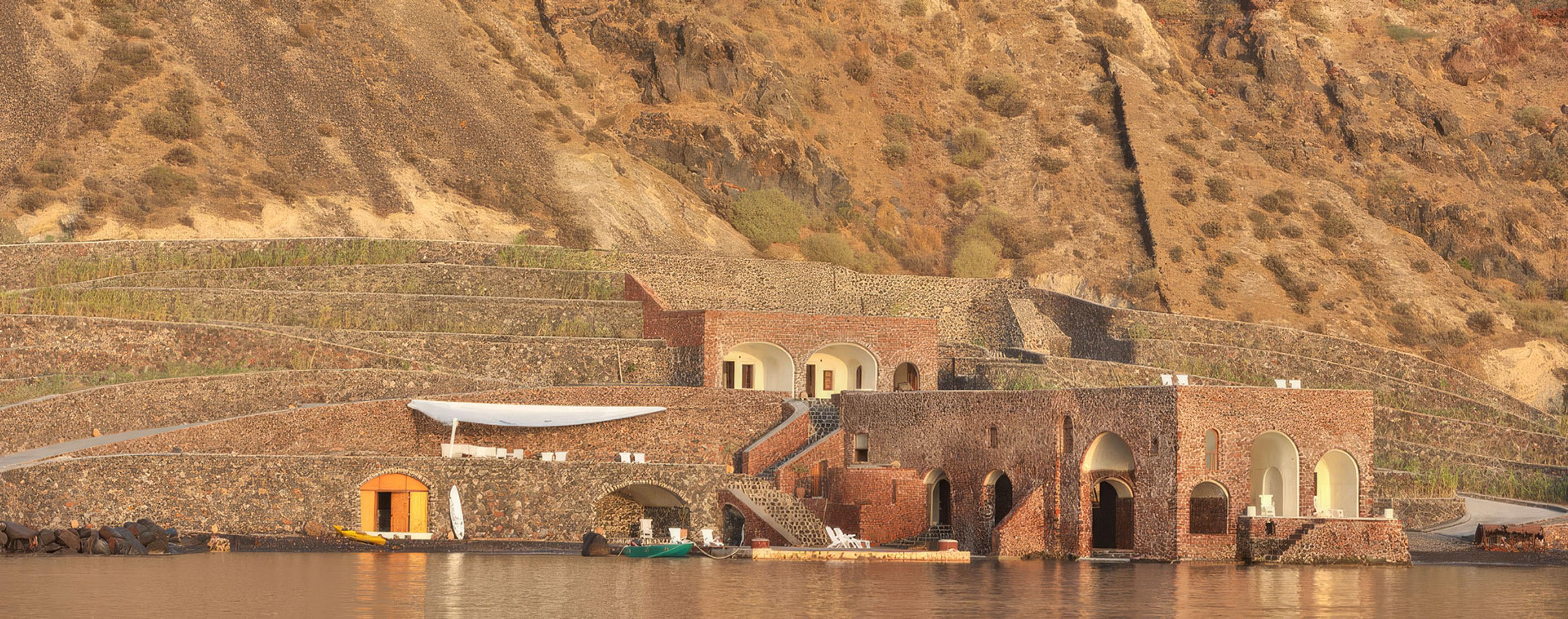 A cluster of stone and whitewashed buildings built into a volcanic cliff above the sea, with boats moored at a stone jetty below.